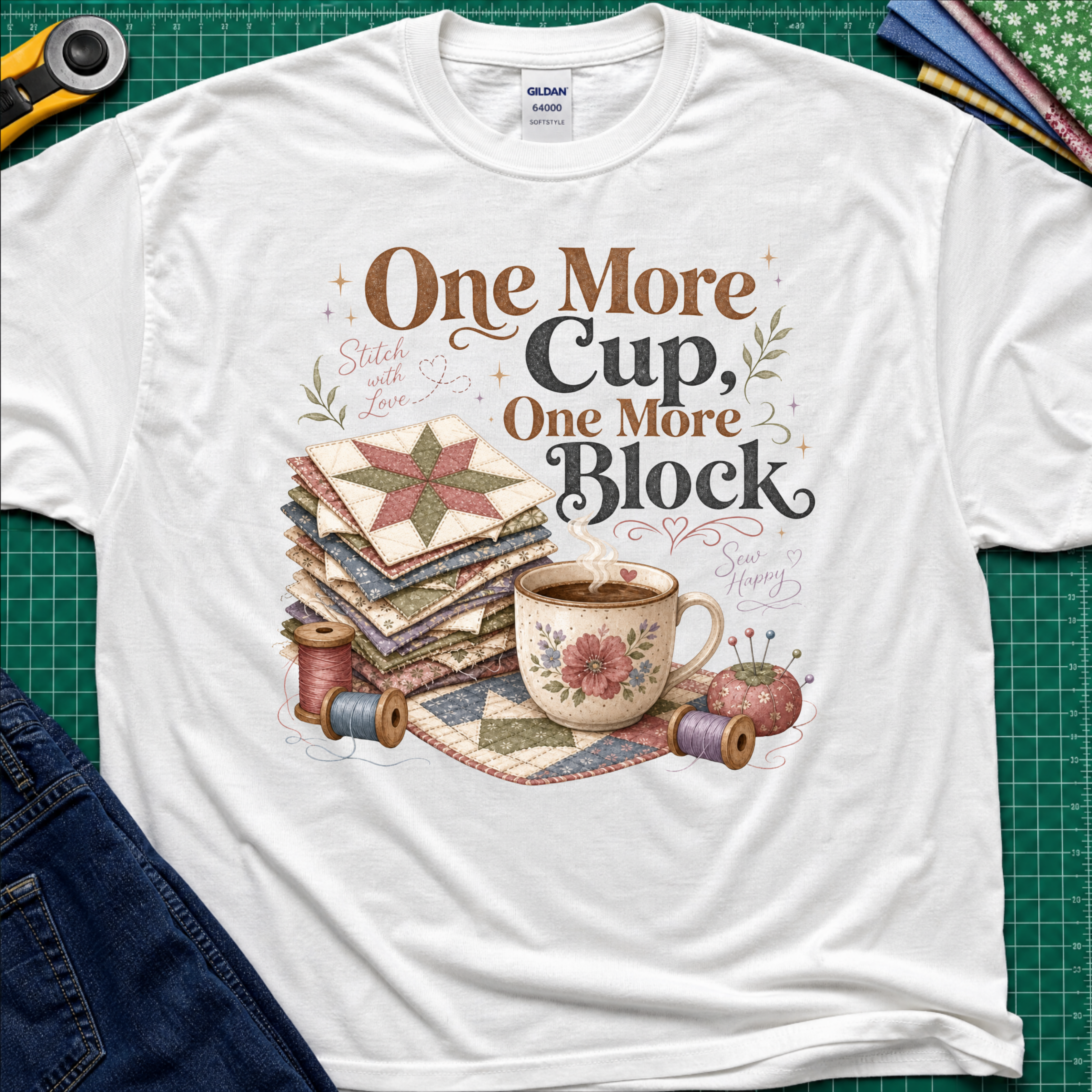 A white crewneck t-shirt that reads 'One More Cup, One More Block' above a floral teacup, a stack of quilt blocks, and thread spools. The shirt is laid flat on a green cutting mat with crafting tools partially visible at the edges.