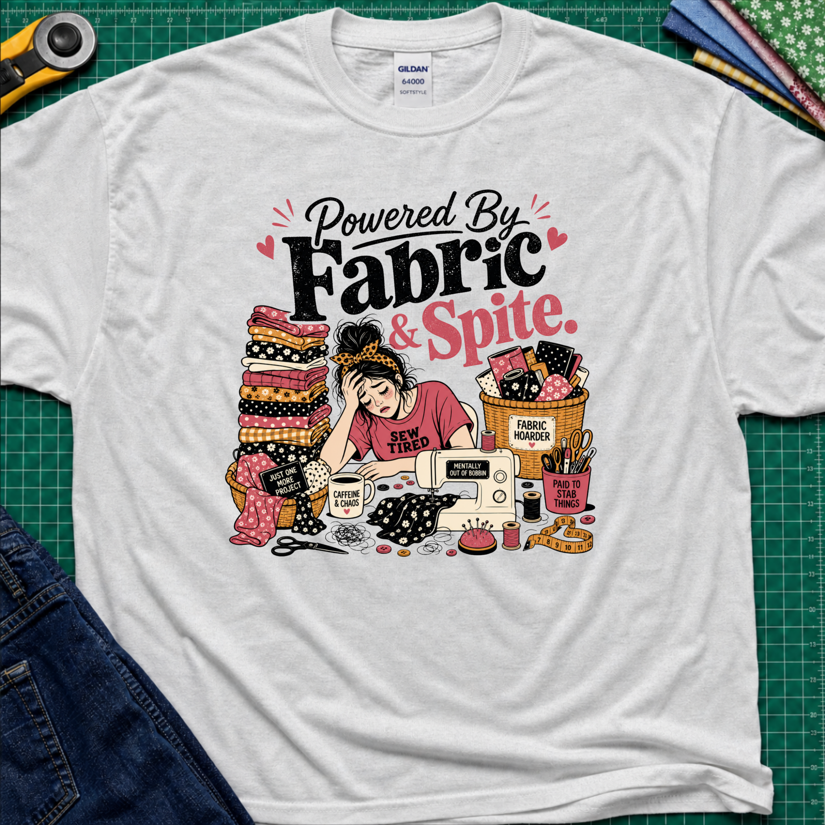 A light grey Gildan t-shirt laid flat on a green cutting mat, featuring a colorful graphic illustration of a sewing woman surrounded by stacks of fabric, a sewing machine, and the text 'Powered By Fabric & Spite.'.