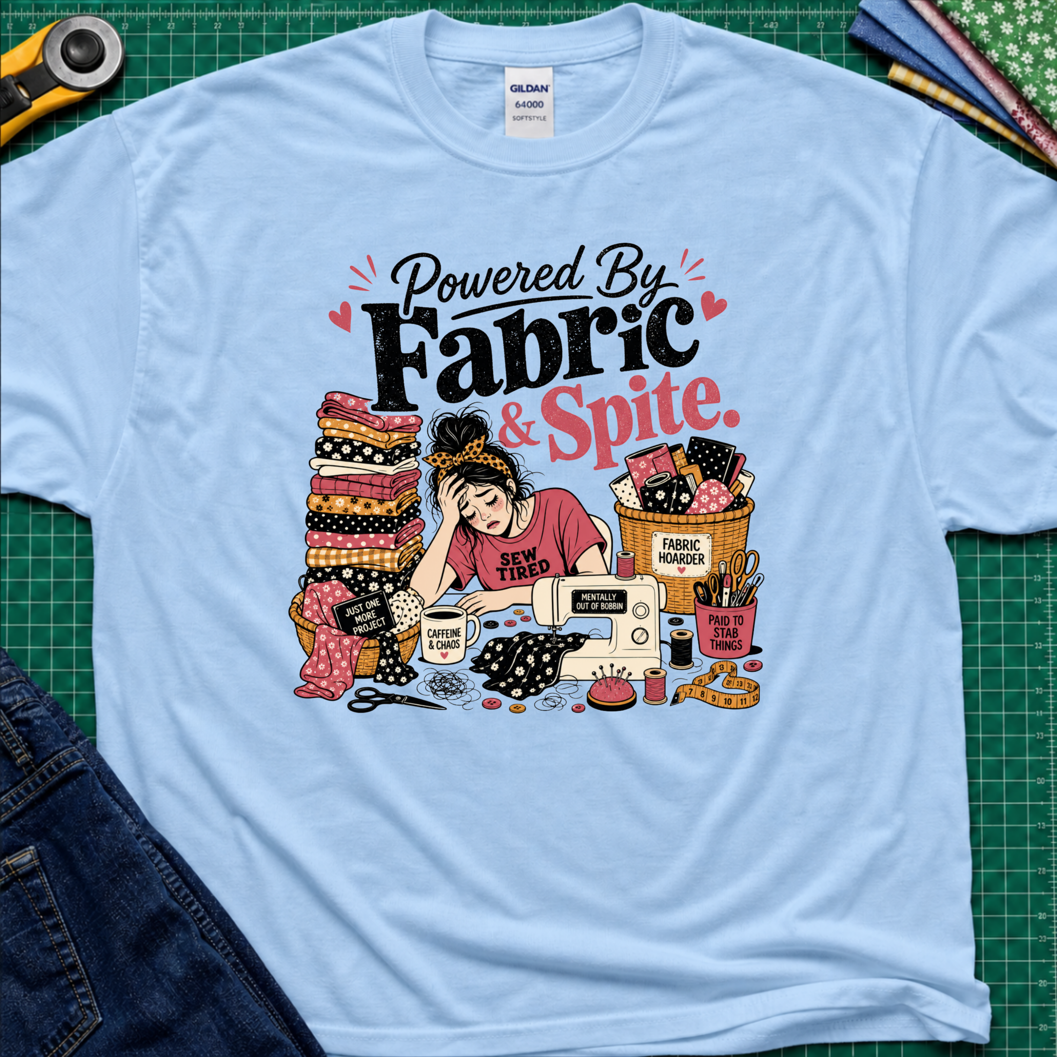 A light blue Gildan t-shirt laid flat on a green cutting mat, featuring a colorful graphic illustration of a sewing woman surrounded by stacks of fabric, a sewing machine, and the text 'Powered By Fabric & Spite.'.