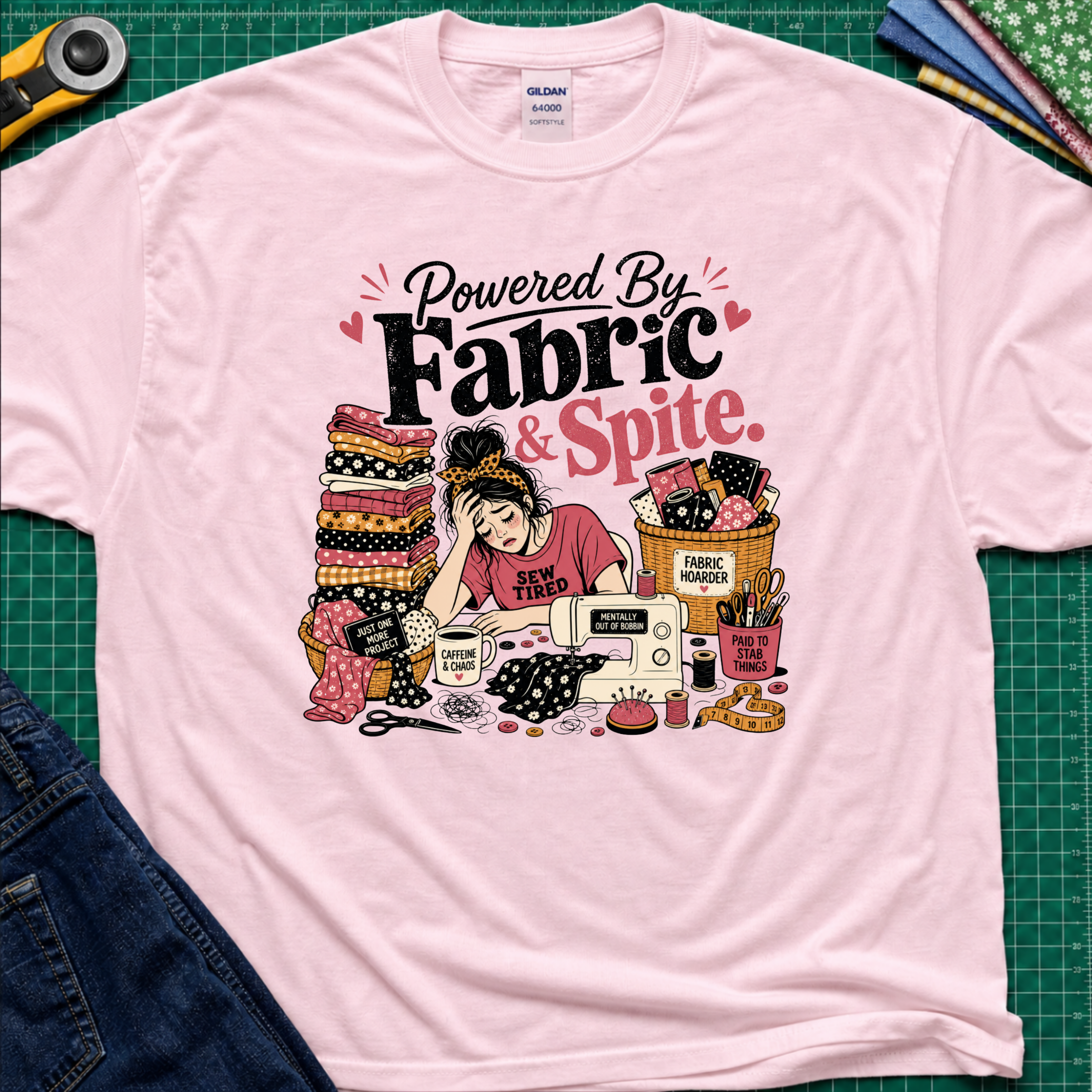 A light pink Gildan t-shirt laid flat on a green cutting mat, featuring a colorful graphic illustration of a sewing woman surrounded by stacks of fabric, a sewing machine, and the text 'Powered By Fabric & Spite.'.