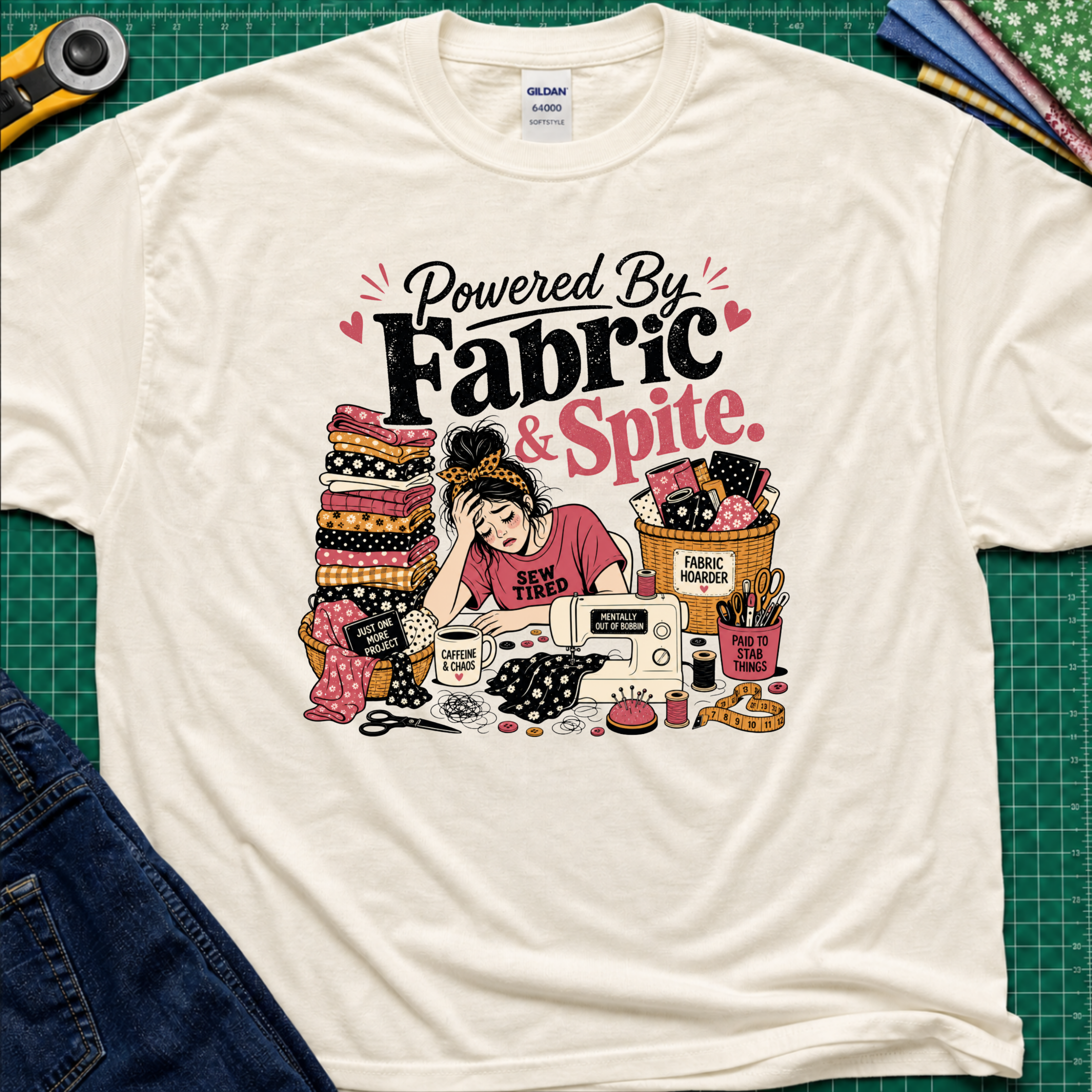 A light cream Gildan t-shirt laid flat on a green cutting mat, featuring a colorful graphic illustration of a sewing woman surrounded by stacks of fabric, a sewing machine, and the text 'Powered By Fabric & Spite.'.