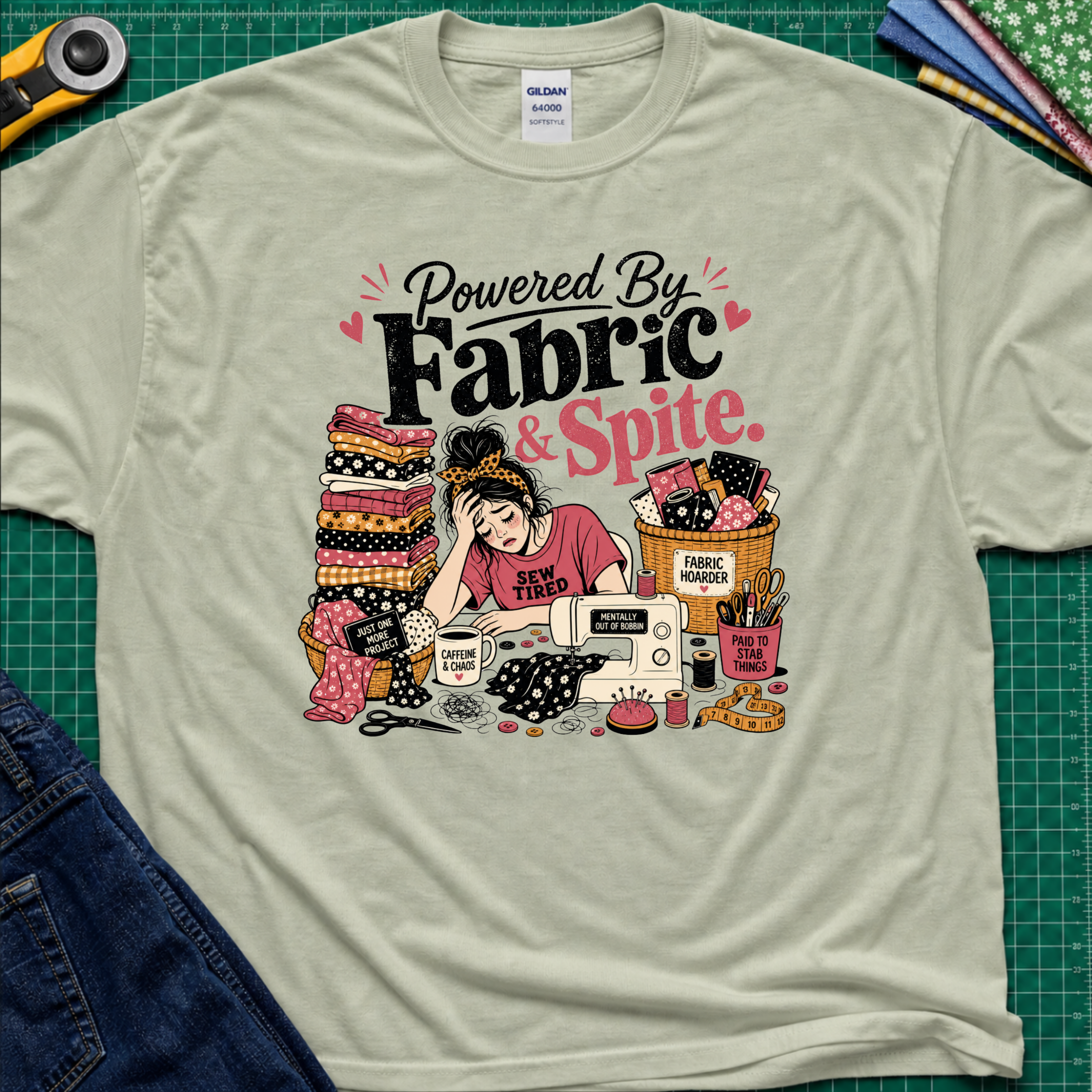 A light sage green Gildan t-shirt laid flat on a green cutting mat, featuring a colorful graphic illustration of a sewing woman surrounded by stacks of fabric, a sewing machine, and the text 'Powered By Fabric & Spite.'.