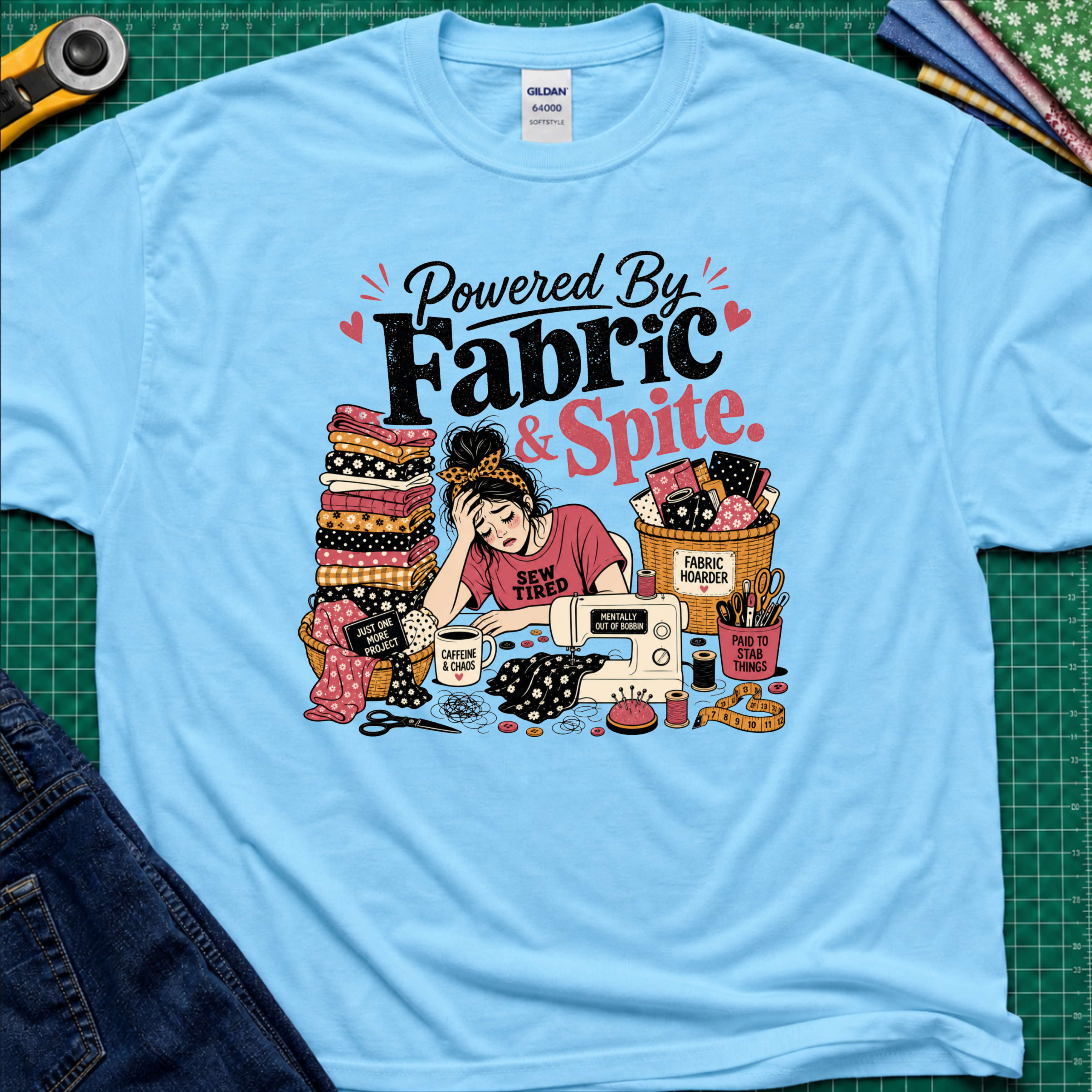A light sky blue Gildan t-shirt laid flat on a green cutting mat, featuring a colorful graphic illustration of a sewing woman surrounded by stacks of fabric, a sewing machine, and the text 'Powered By Fabric & Spite.'.