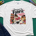 A white Gildan t-shirt laid flat on a green cutting mat, featuring a colorful graphic illustration of a sewing woman surrounded by stacks of fabric, a sewing machine, and the text 'Powered By Fabric & Spite.'.