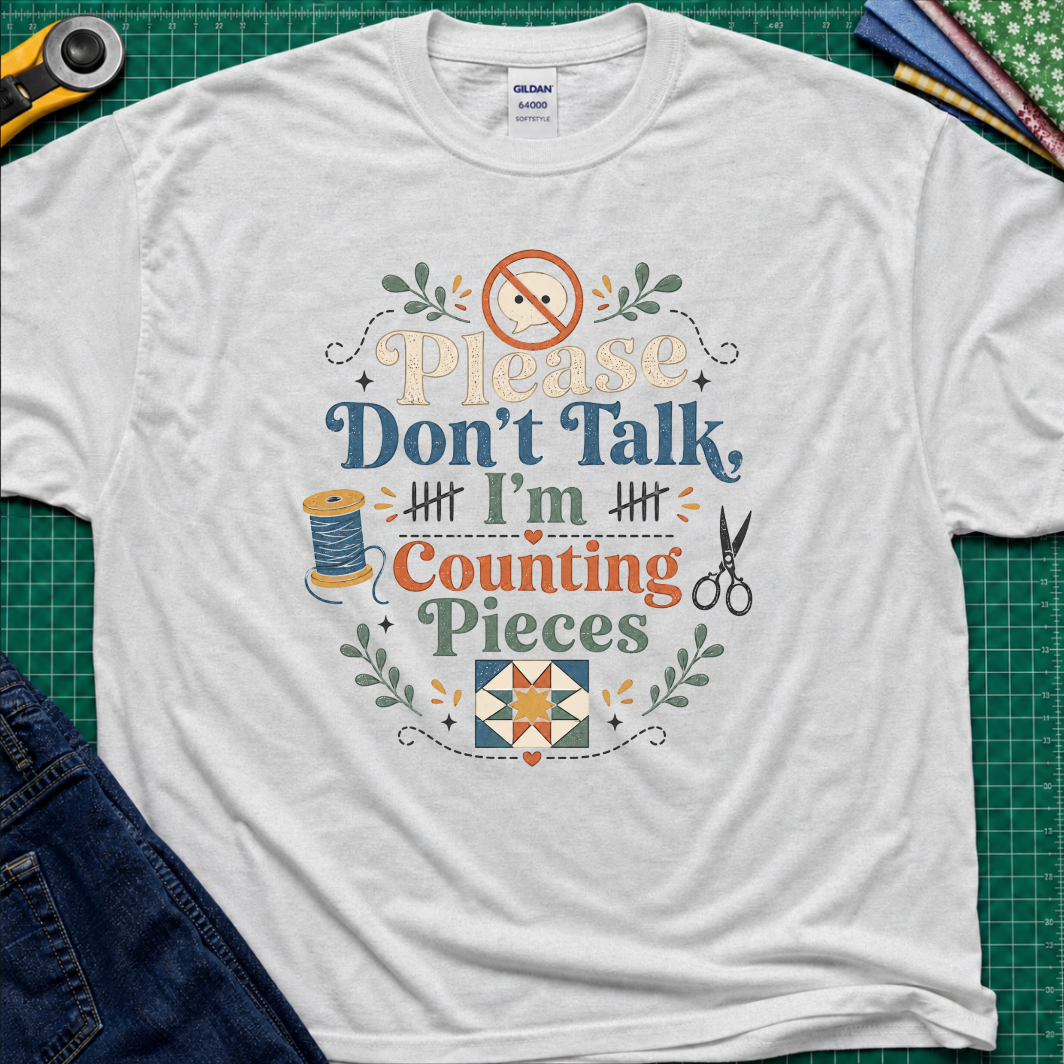 A light grey Gildan t-shirt with a colorful graphic that reads 'Please Don't Talk I'm Counting Pieces' with sewing-themed illustrations, laid flat on a green cutting mat with crafting tools around it.