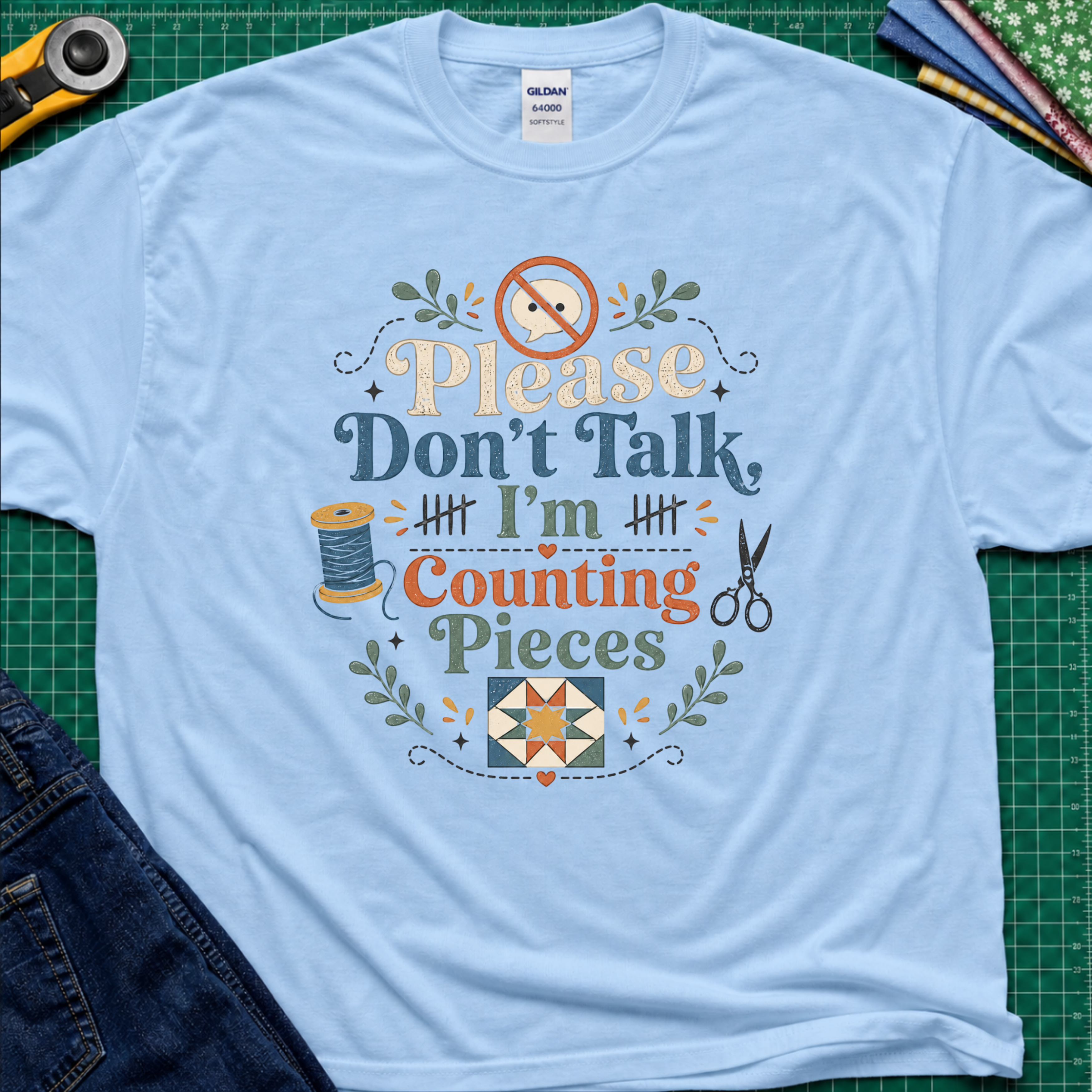 A light blue Gildan t-shirt with a colorful graphic that reads 'Please Don't Talk I'm Counting Pieces' with sewing-themed illustrations, laid flat on a green cutting mat with crafting tools around it.