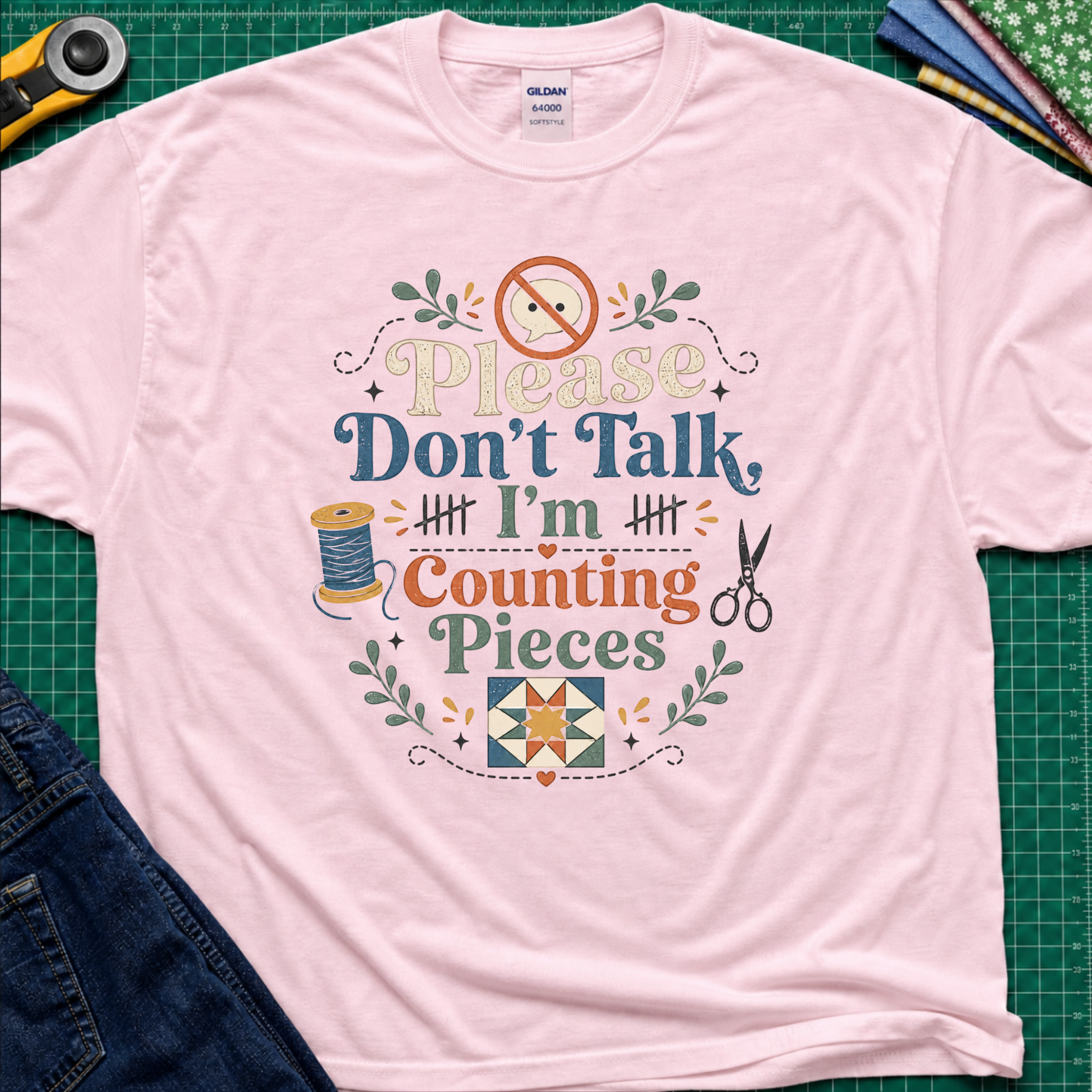 A light pink Gildan t-shirt with a colorful graphic that reads 'Please Don't Talk I'm Counting Pieces' with sewing-themed illustrations, laid flat on a green cutting mat with crafting tools around it.
