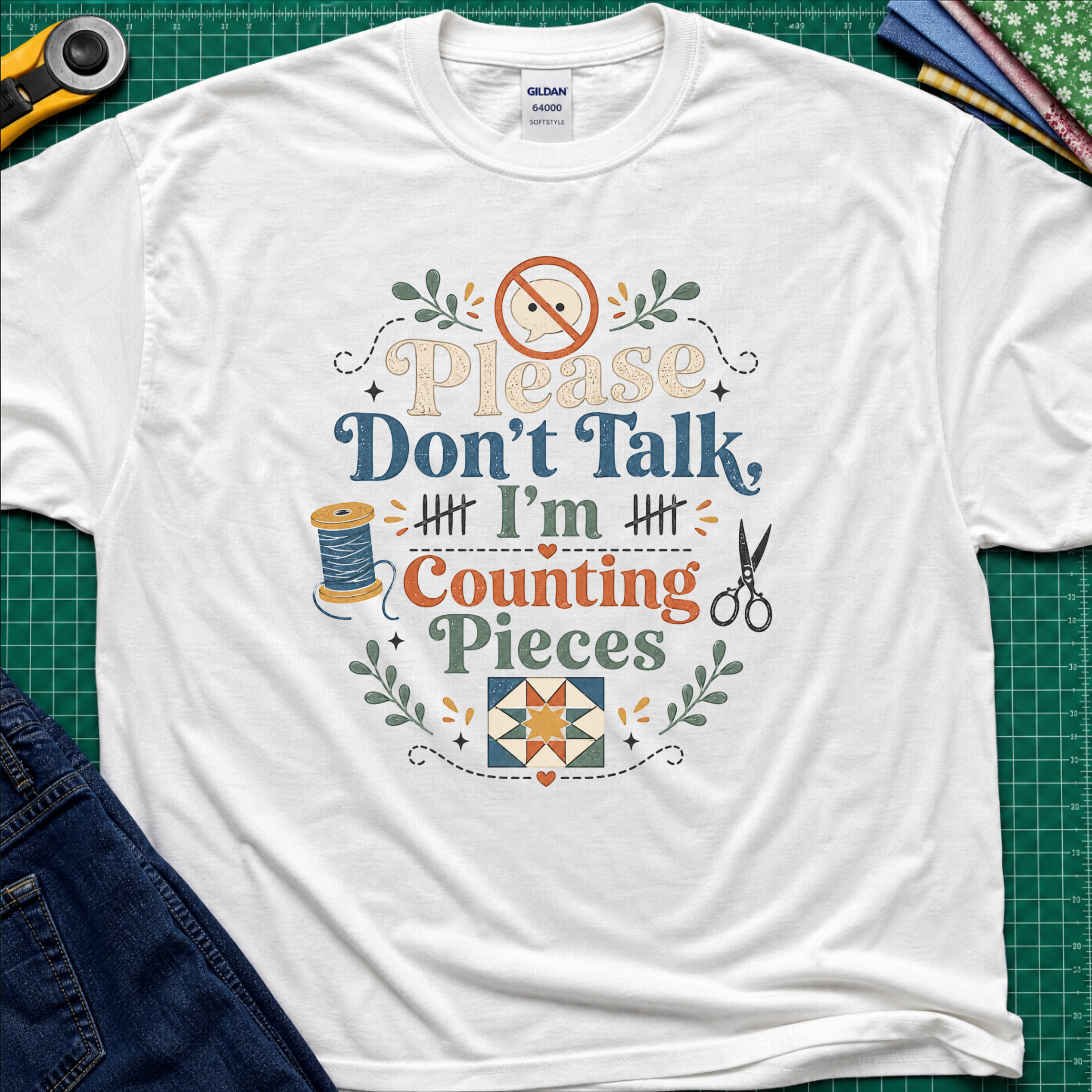 A white Gildan t-shirt with a colorful graphic that reads 'Please Don't Talk I'm Counting Pieces' with sewing-themed illustrations, laid flat on a green cutting mat with crafting tools around it.