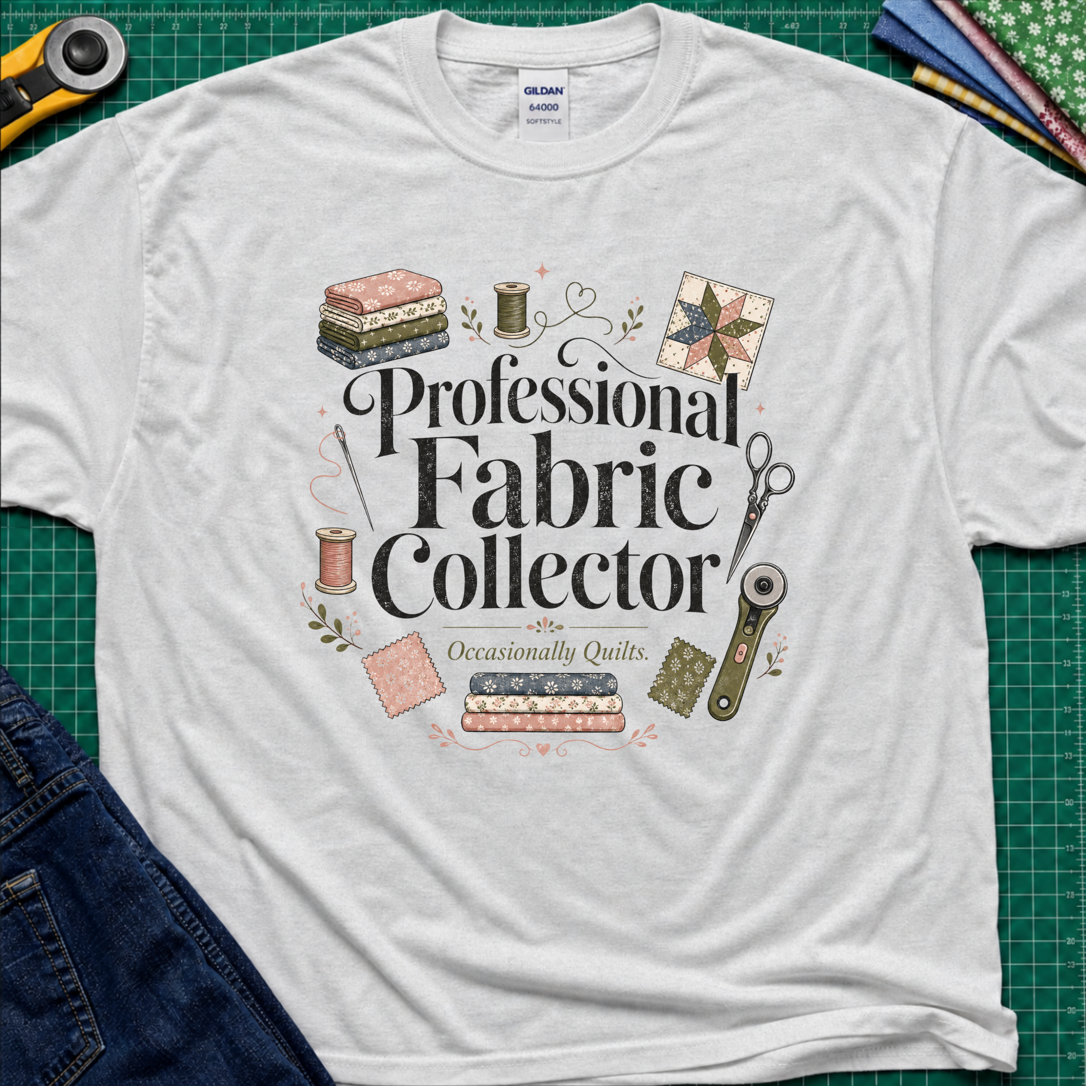 A light grey Gildan t-shirt laid flat on a green cutting mat, surrounded by sewing props including a rotary cutter, fabric, thread, scissors, and denim jeans. The shirt features the text 'Professional Fabric Collector' with 'Occasionally Quits.