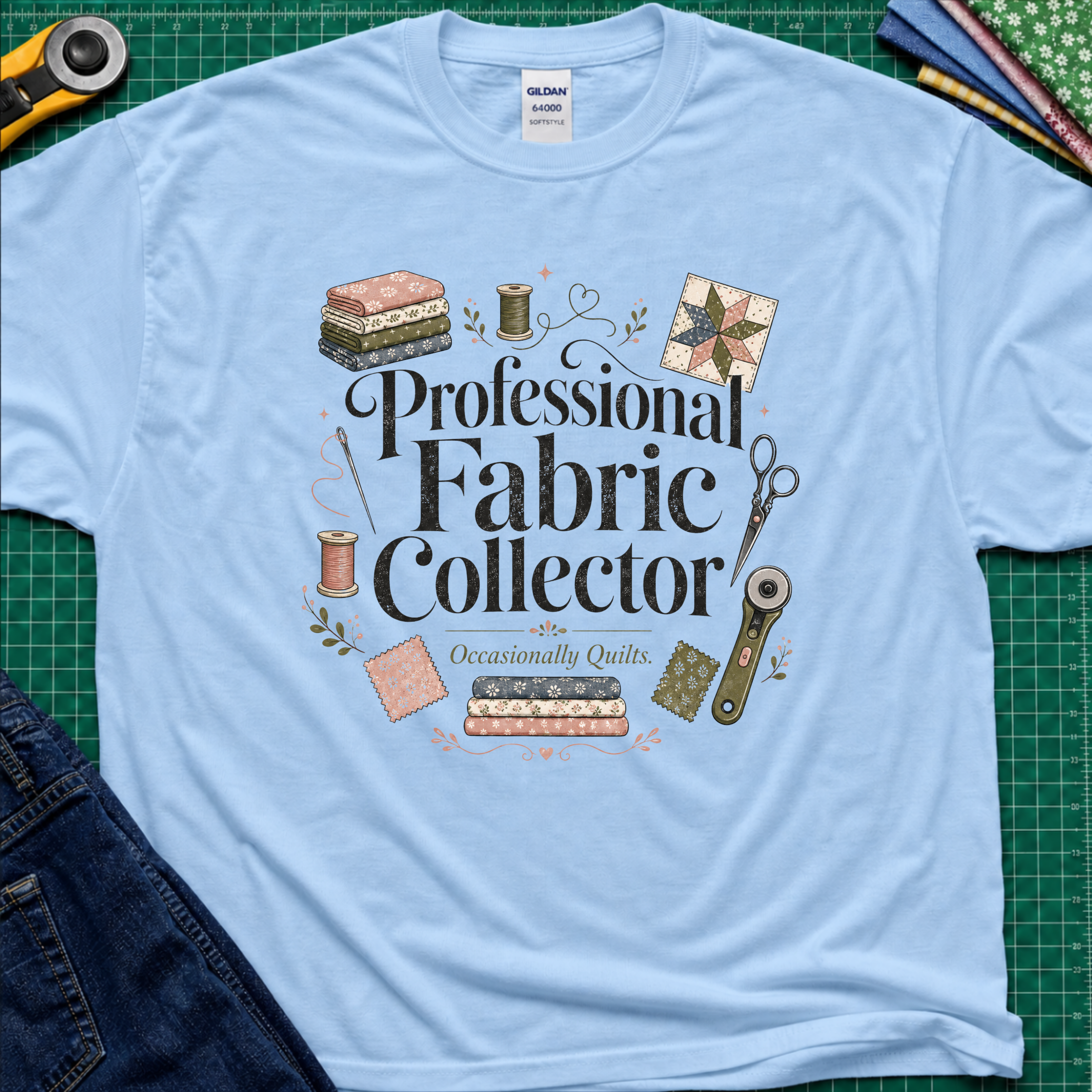 A light blue Gildan t-shirt laid flat on a green cutting mat, surrounded by sewing props including a rotary cutter, fabric, thread, scissors, and denim jeans. The shirt features the text 'Professional Fabric Collector' with 'Occasionally Quits.