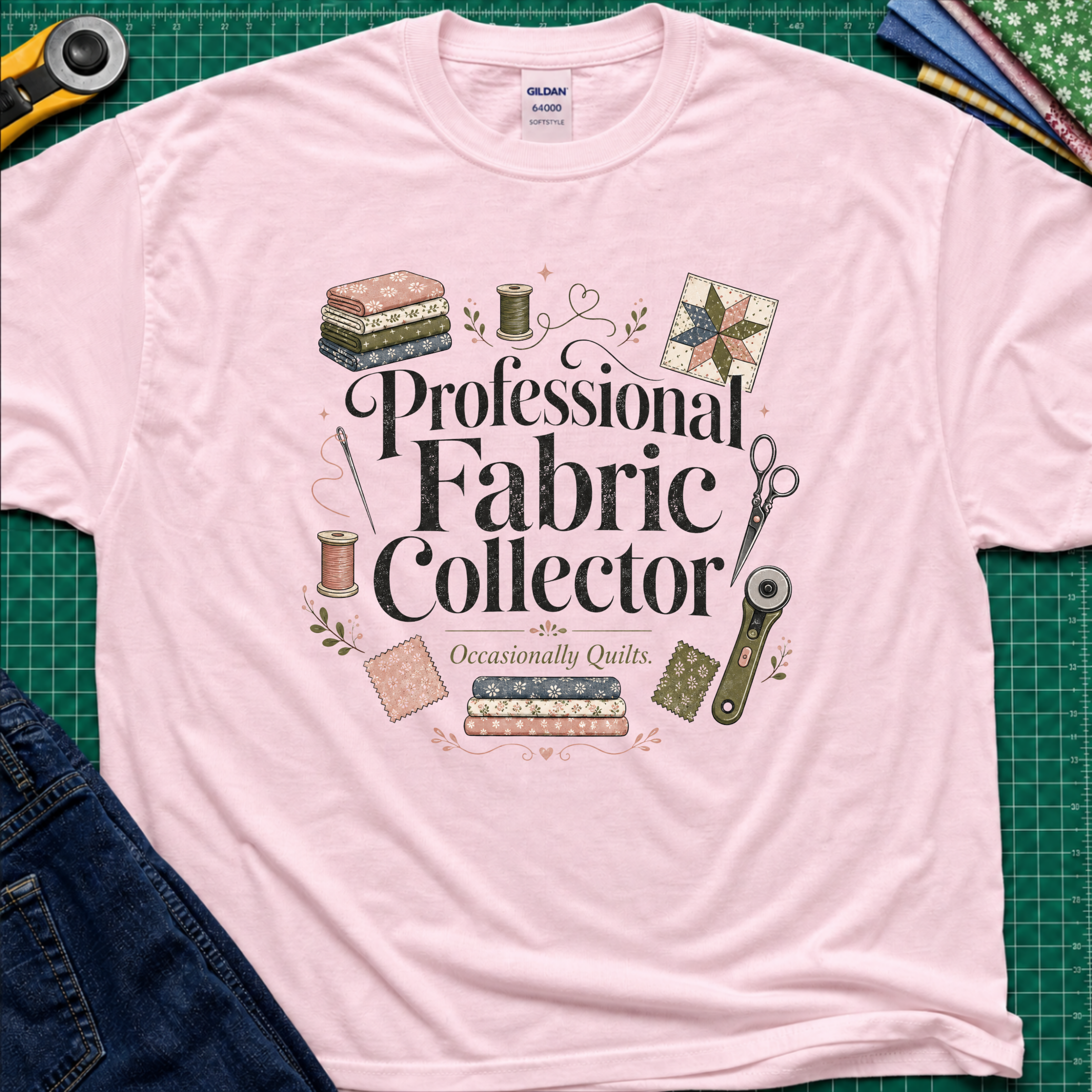 A light pink Gildan t-shirt laid flat on a green cutting mat, surrounded by sewing props including a rotary cutter, fabric, thread, scissors, and denim jeans. The shirt features the text 'Professional Fabric Collector' with 'Occasionally Quits.
