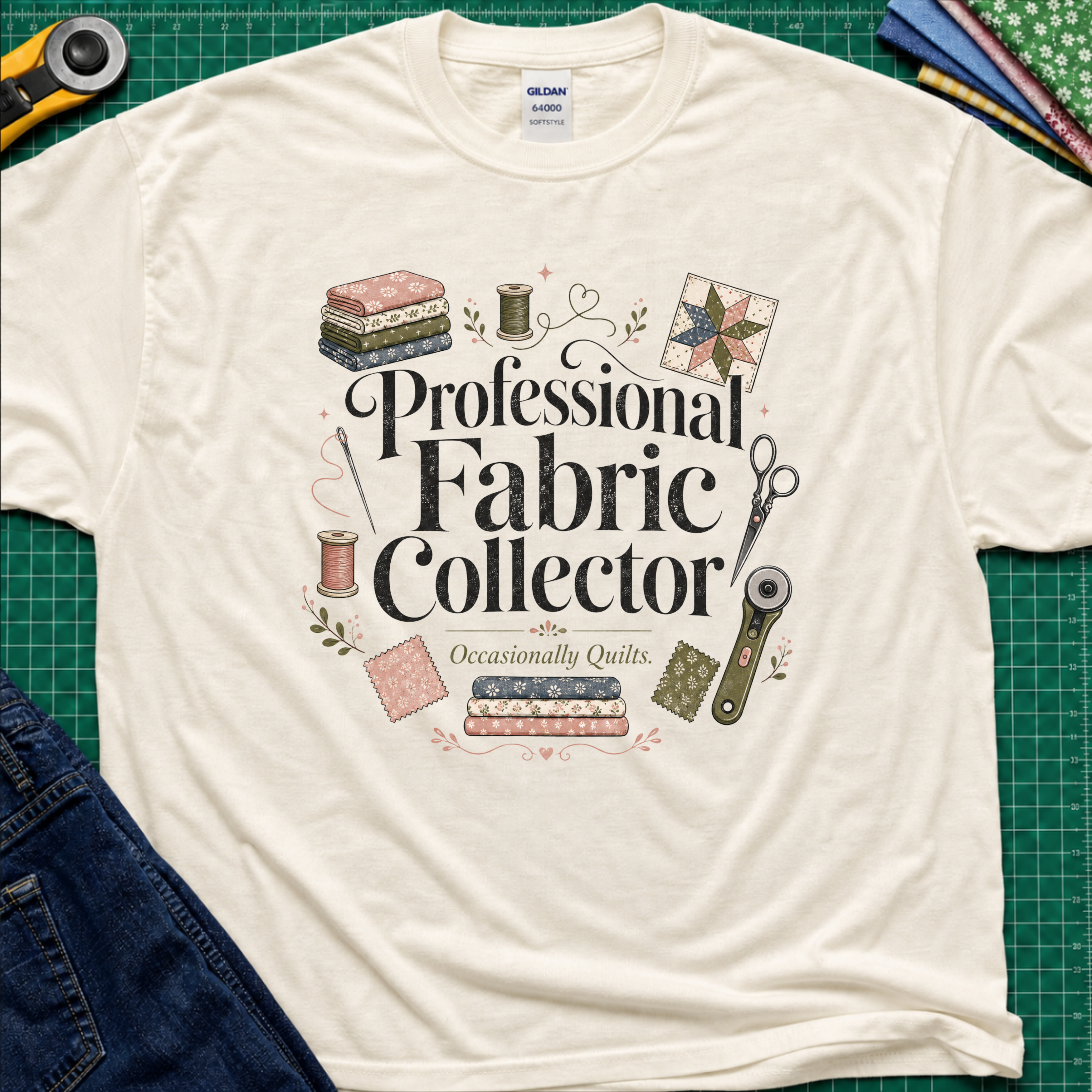 A light cream Gildan t-shirt laid flat on a green cutting mat, surrounded by sewing props including a rotary cutter, fabric, thread, scissors, and denim jeans. The shirt features the text 'Professional Fabric Collector' with 'Occasionally Quits.