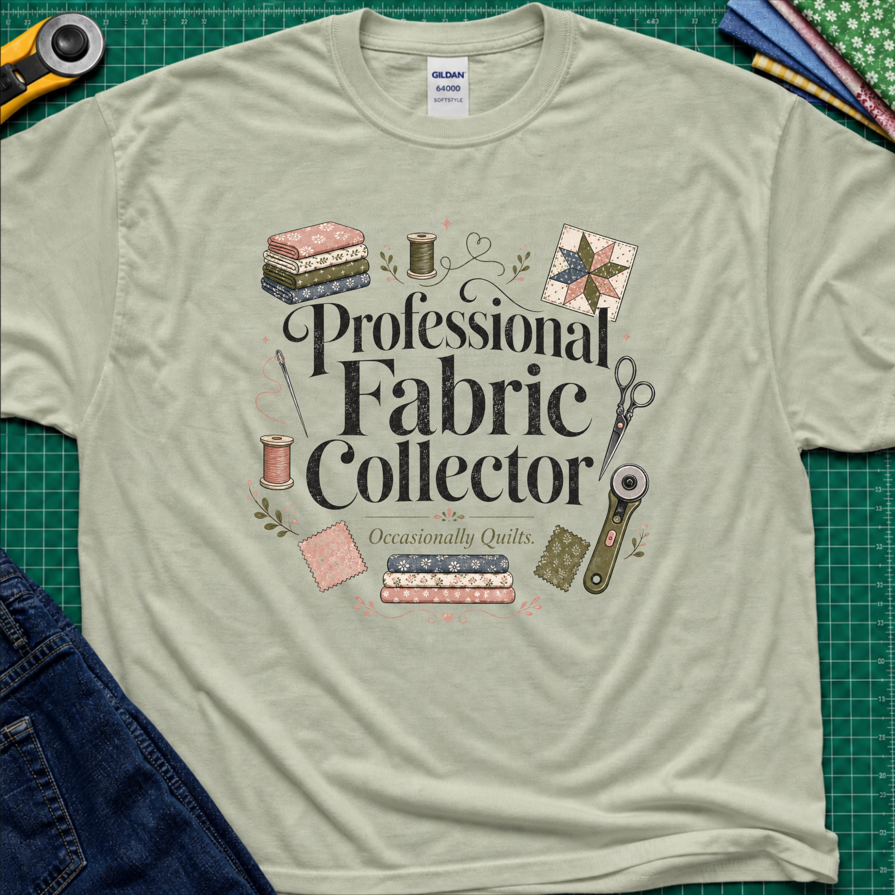 A light sage green Gildan t-shirt laid flat on a green cutting mat, surrounded by sewing props including a rotary cutter, fabric, thread, scissors, and denim jeans. The shirt features the text 'Professional Fabric Collector' with 'Occasionally Quits.