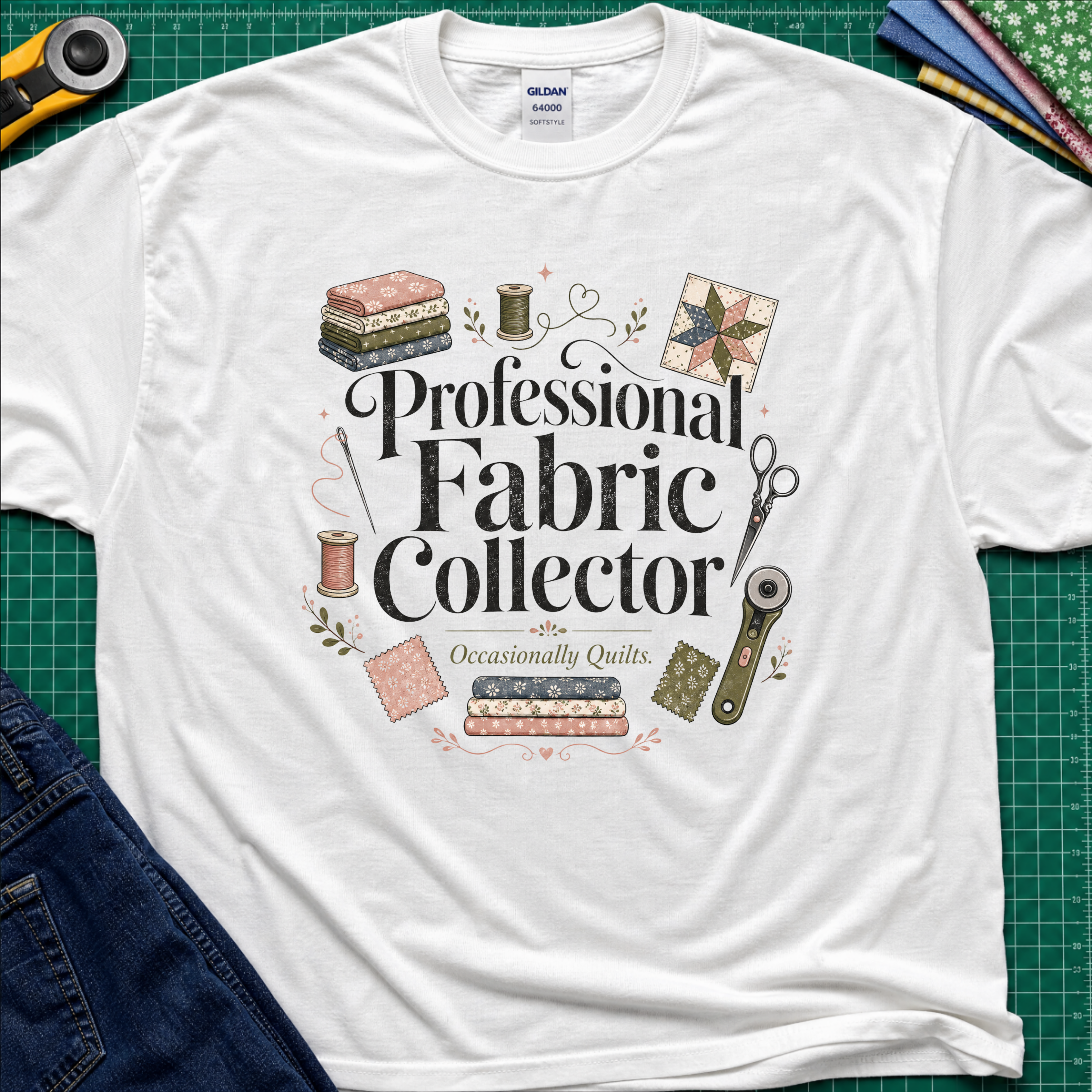 A white Gildan t-shirt laid flat on a green cutting mat, surrounded by sewing props including a rotary cutter, fabric, thread, scissors, and denim jeans. The shirt features the text 'Professional Fabric Collector' with 'Occasionally Quits.