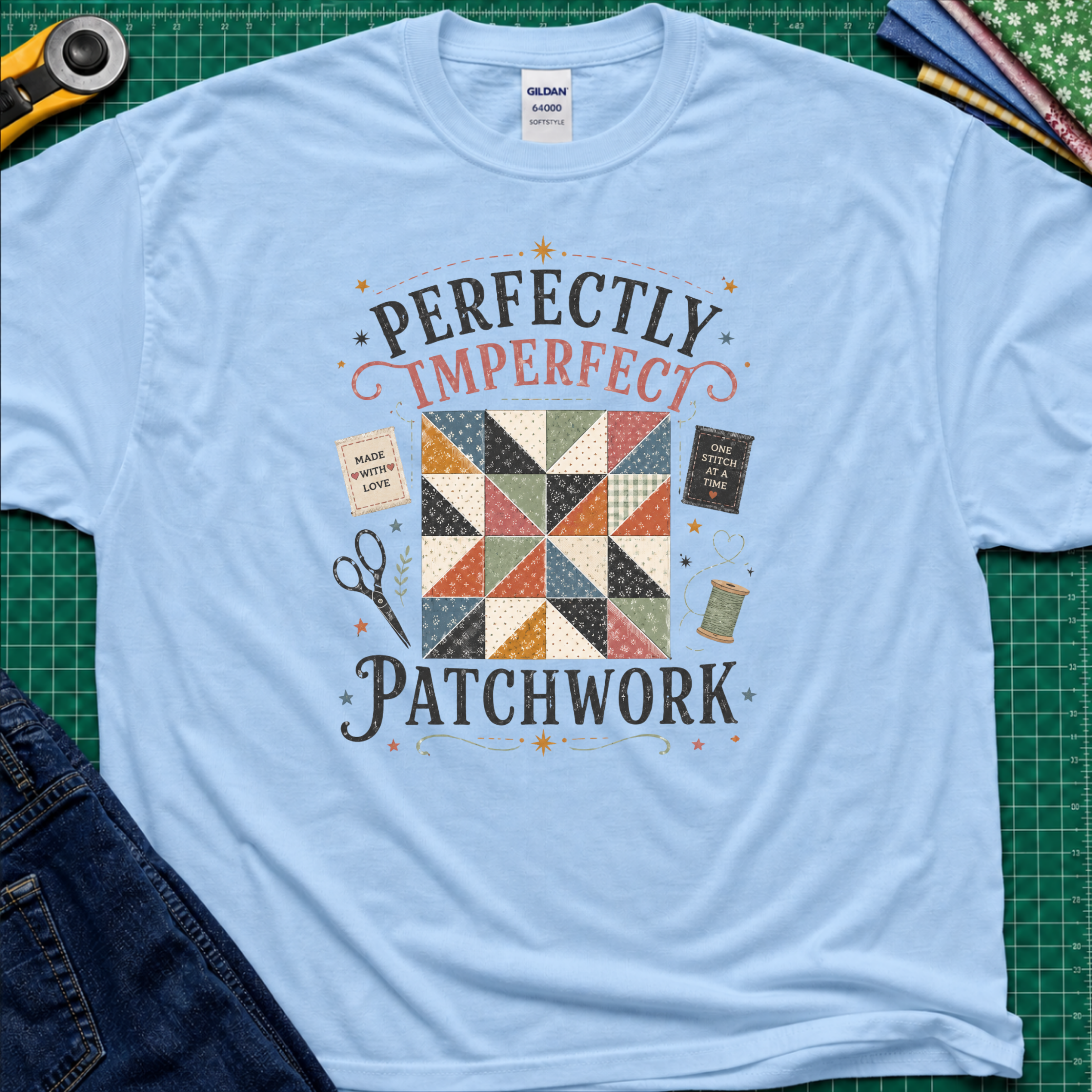 A light blue Gildan t-shirt laid flat on a green cutting mat, featuring a patchwork quilt graphic with the words 'PERFECTLY IMPERFECT PATCHWORK.' Surrounding props include a rotary cutter, fabric, scissors, and jeans.