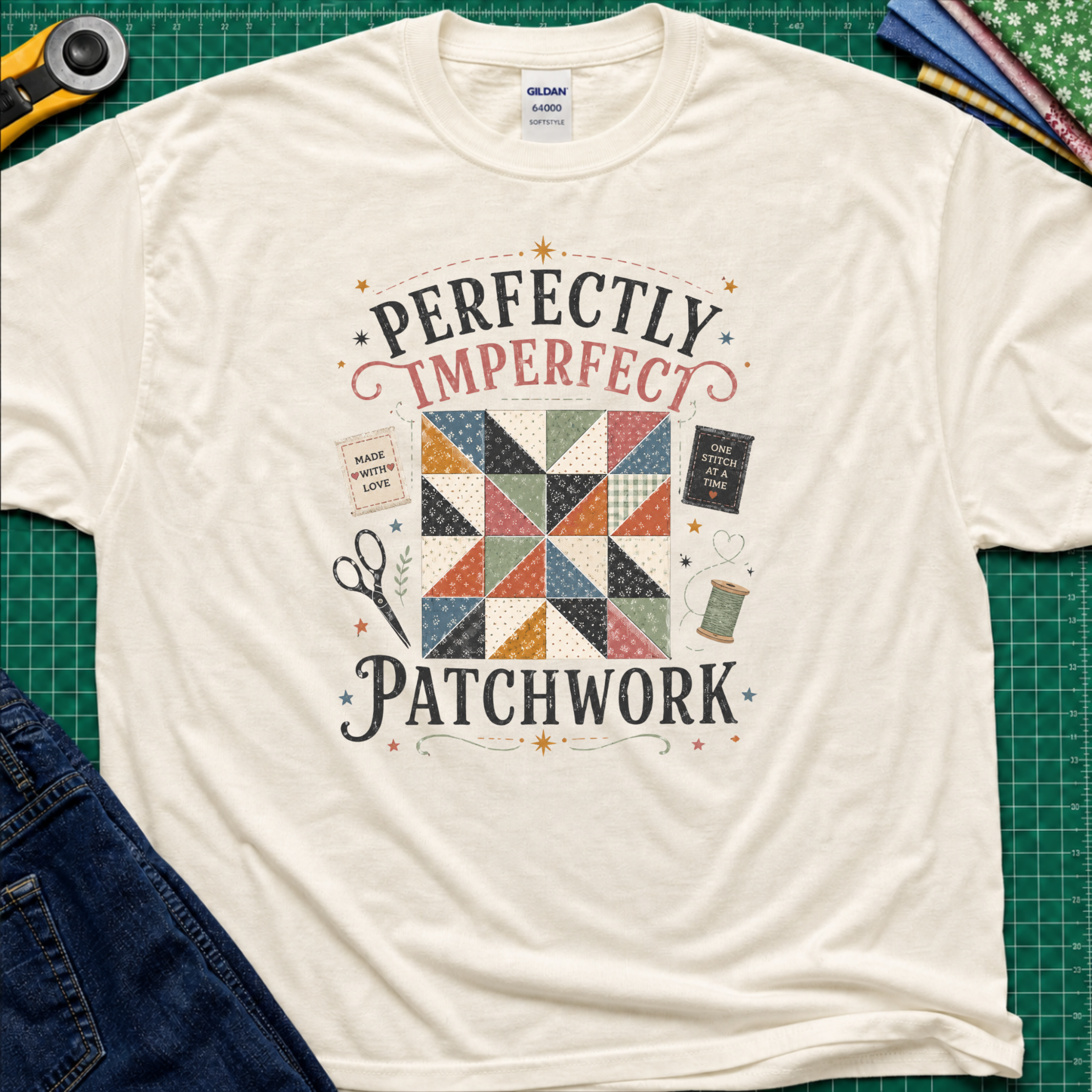 A light cream Gildan t-shirt laid flat on a green cutting mat, featuring a patchwork quilt graphic with the words 'PERFECTLY IMPERFECT PATCHWORK.' Surrounding props include a rotary cutter, fabric, scissors, and jeans.