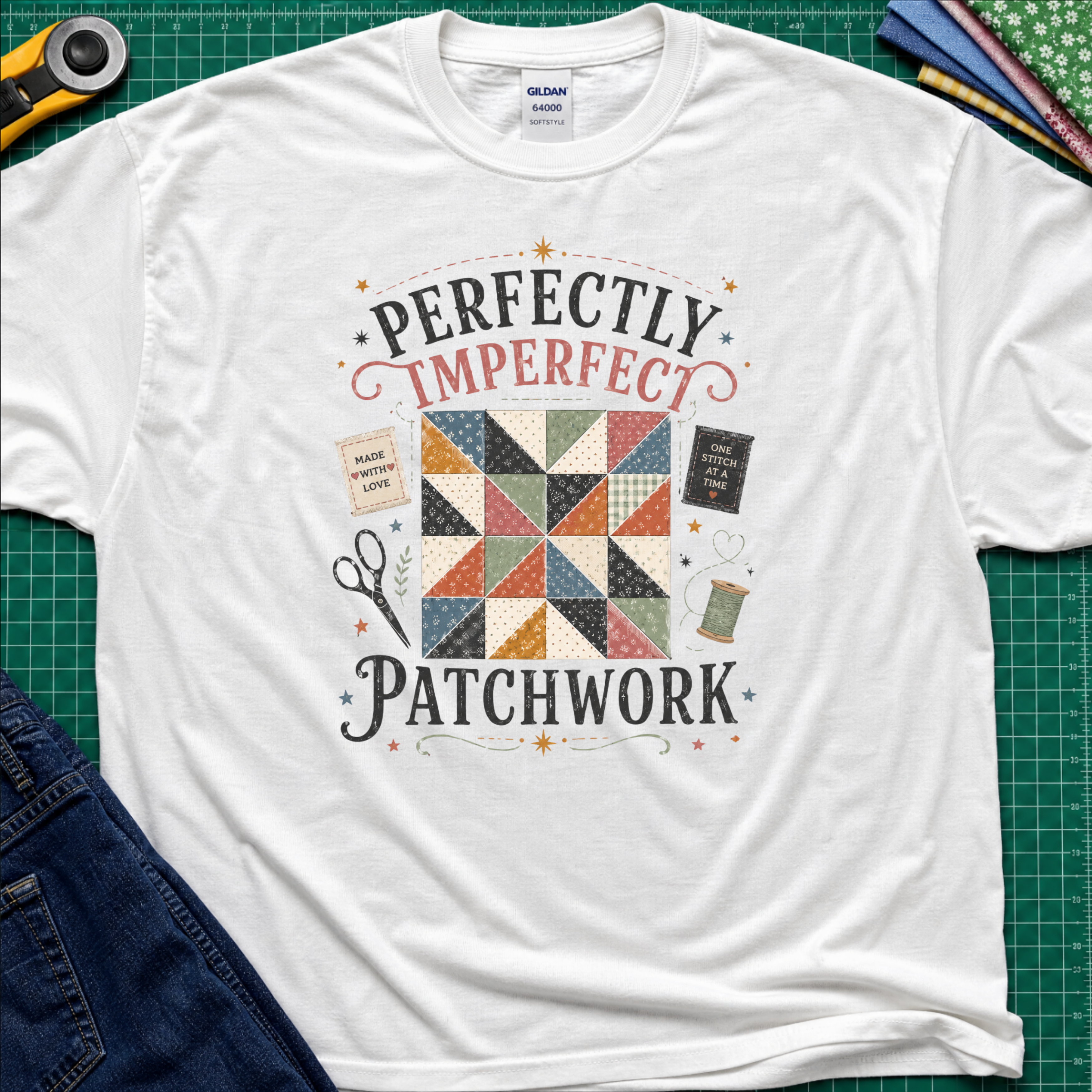 A white Gildan t-shirt laid flat on a green cutting mat, featuring a patchwork quilt graphic with the words 'PERFECTLY IMPERFECT PATCHWORK.' Surrounding props include a rotary cutter, fabric, scissors, and jeans.