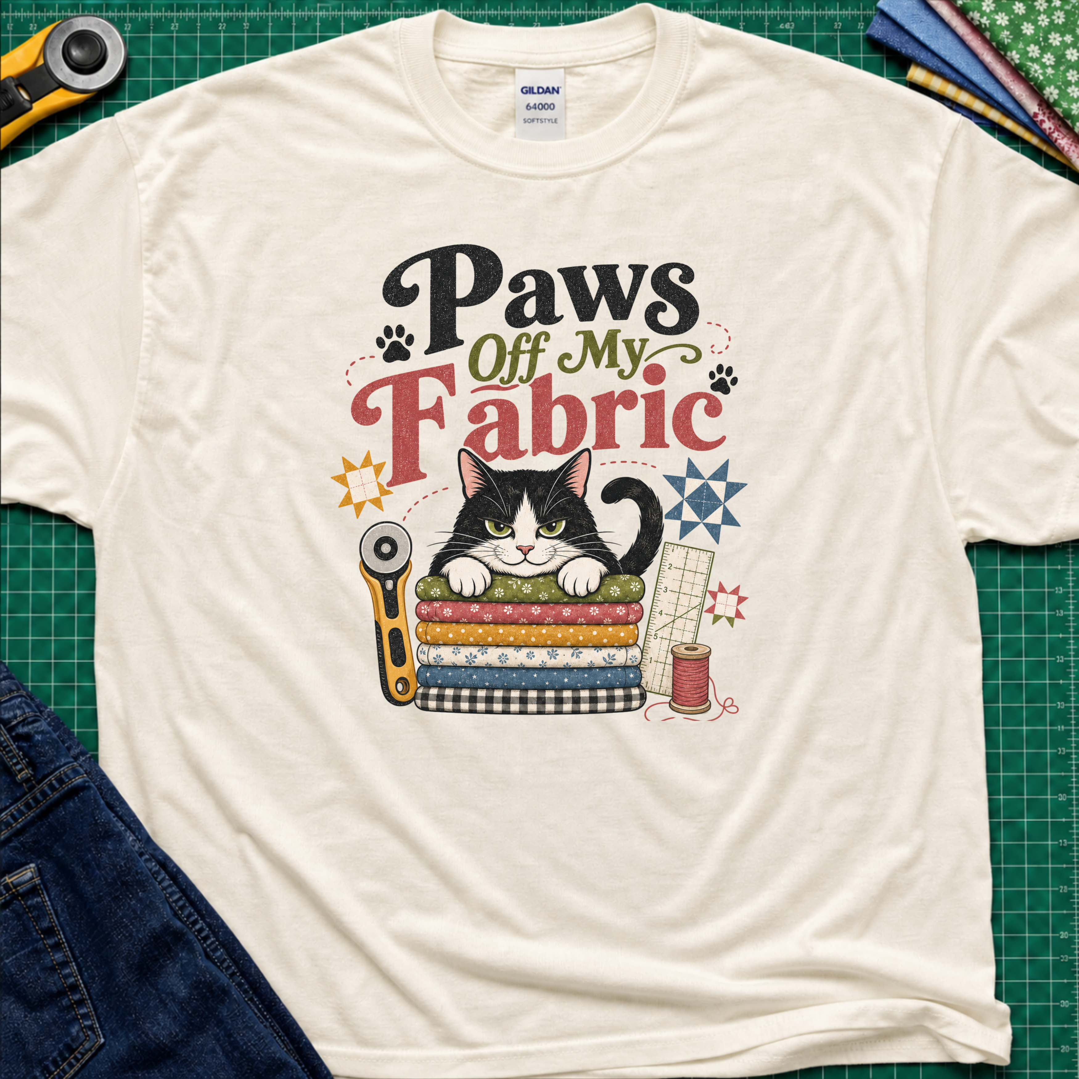 A light cream Gildan t-shirt laid flat on a green cutting mat, featuring a colorful graphic of a black and white cat behind stacked fabrics with the text 'Paws Off My Fabric'. Quilting tools and fabrics are styled around the shirt.