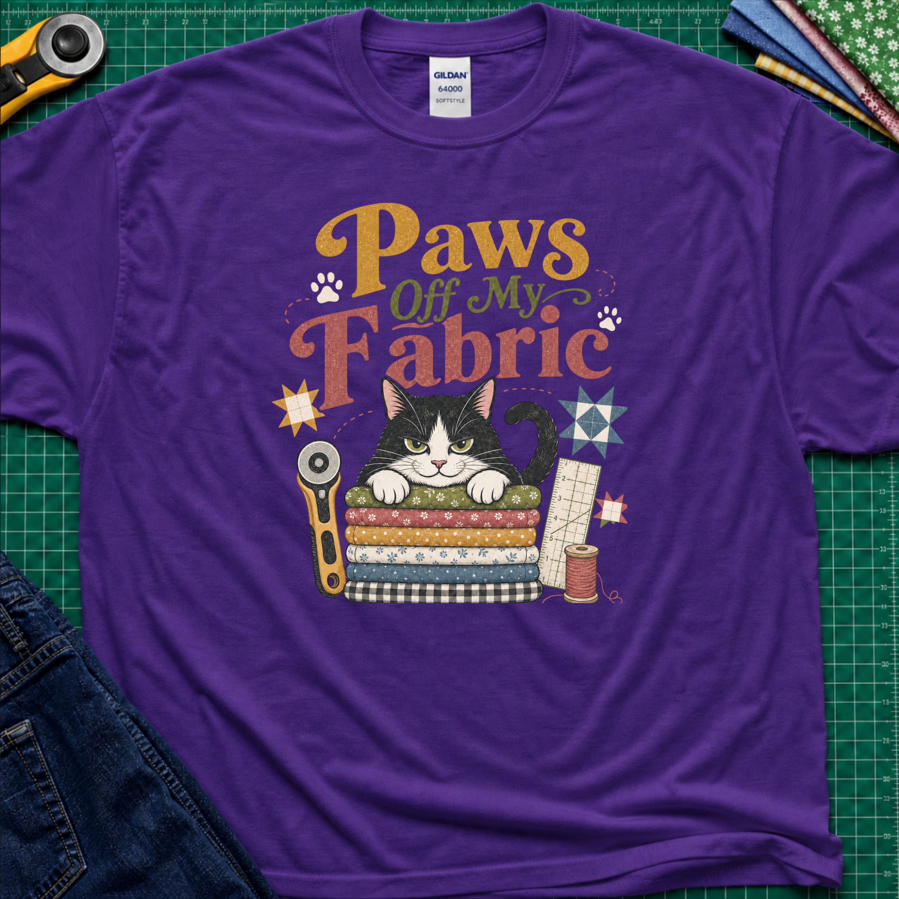 A purple Gildan t-shirt laid flat on a green cutting mat, featuring a colorful graphic of a black and white cat behind stacked fabrics with the text 'Paws Off My Fabric'. Quilting tools and fabrics are styled around the shirt.