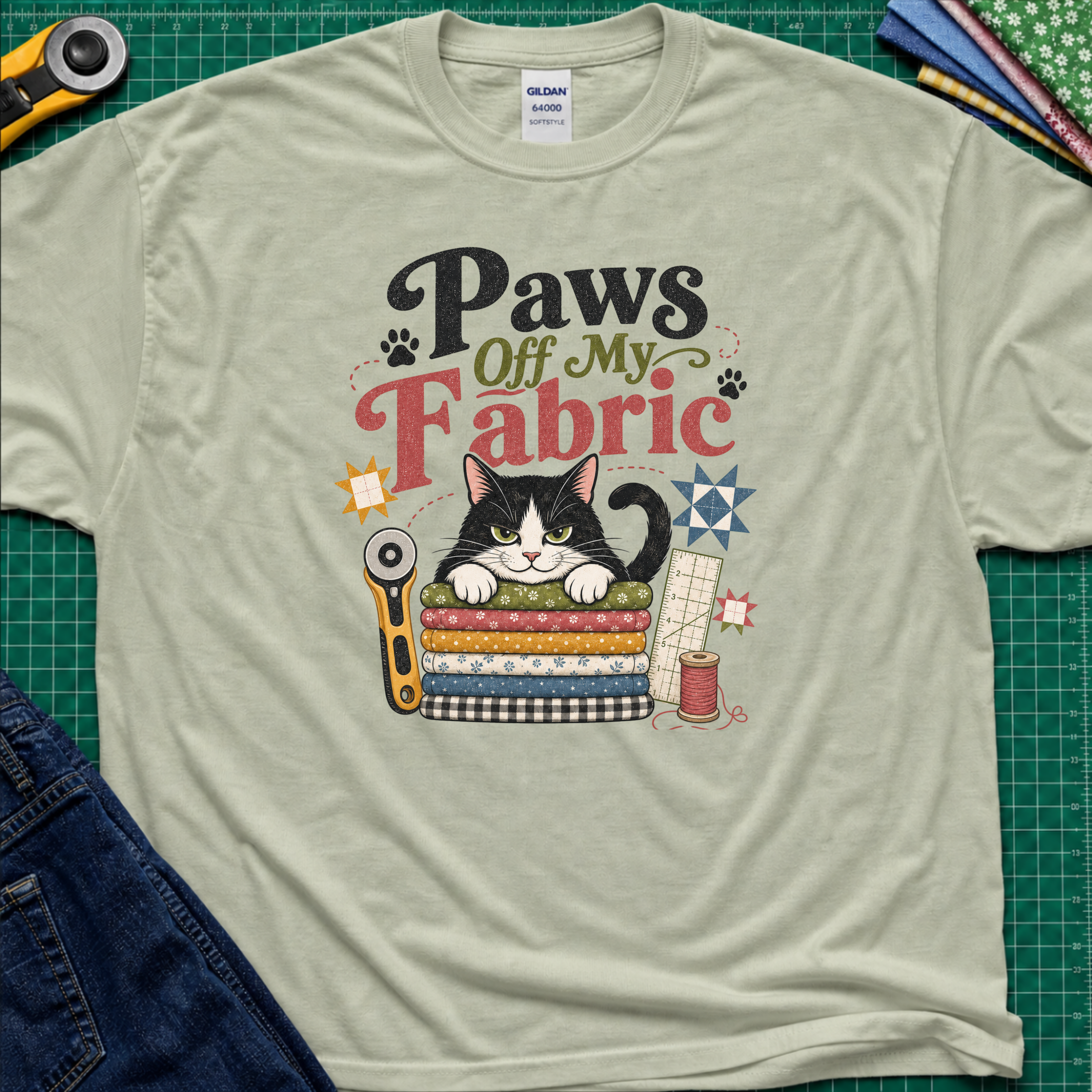 A light sage green Gildan t-shirt laid flat on a green cutting mat, featuring a colorful graphic of a black and white cat behind stacked fabrics with the text 'Paws Off My Fabric'. Quilting tools and fabrics are styled around the shirt.