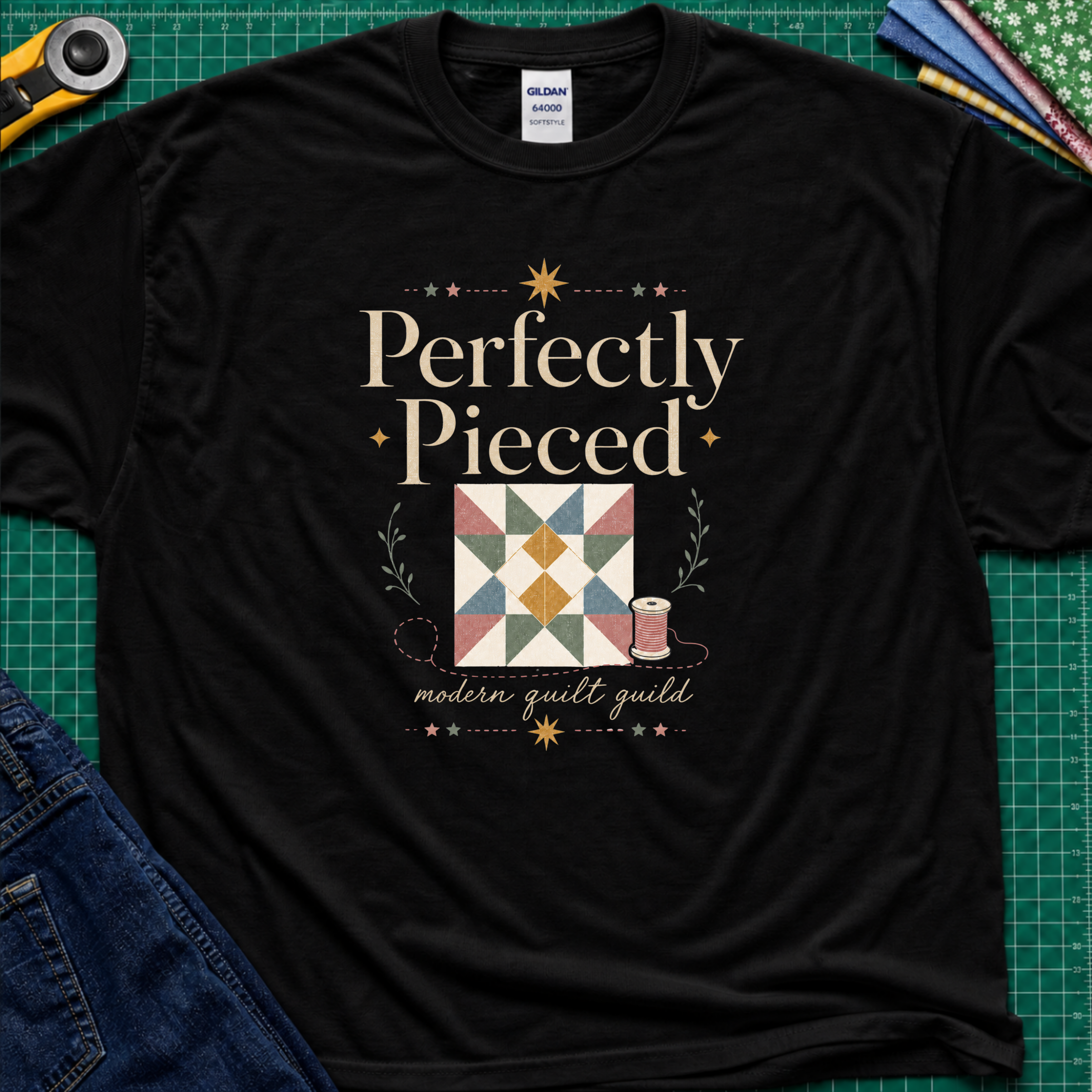 A black Gildan t-shirt with a quilt-themed graphic reading 'Perfectly Pieced' and 'modern quilt guild' around a patchwork block with thread spool, stars and leaves. The shirt lays flat on a green cutting mat with a rotary cutter, fabric stack and blue jeans partially visible in the corners.
