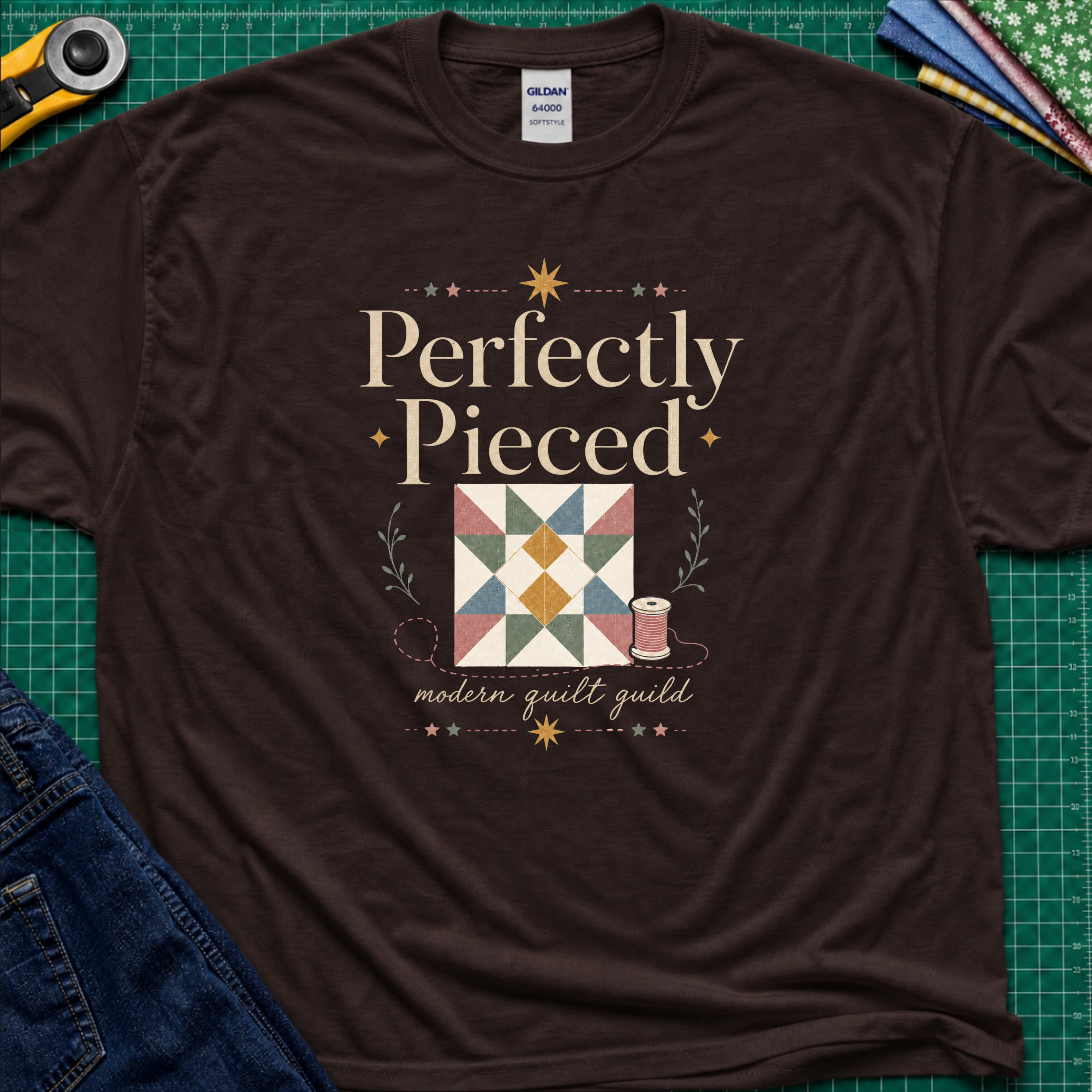 A dark chocolate Gildan t-shirt with a quilt-themed graphic reading 'Perfectly Pieced' and 'modern quilt guild' around a patchwork block with thread spool, stars and leaves. The shirt lays flat on a green cutting mat with a rotary cutter, fabric stack and blue jeans partially visible in the corners.