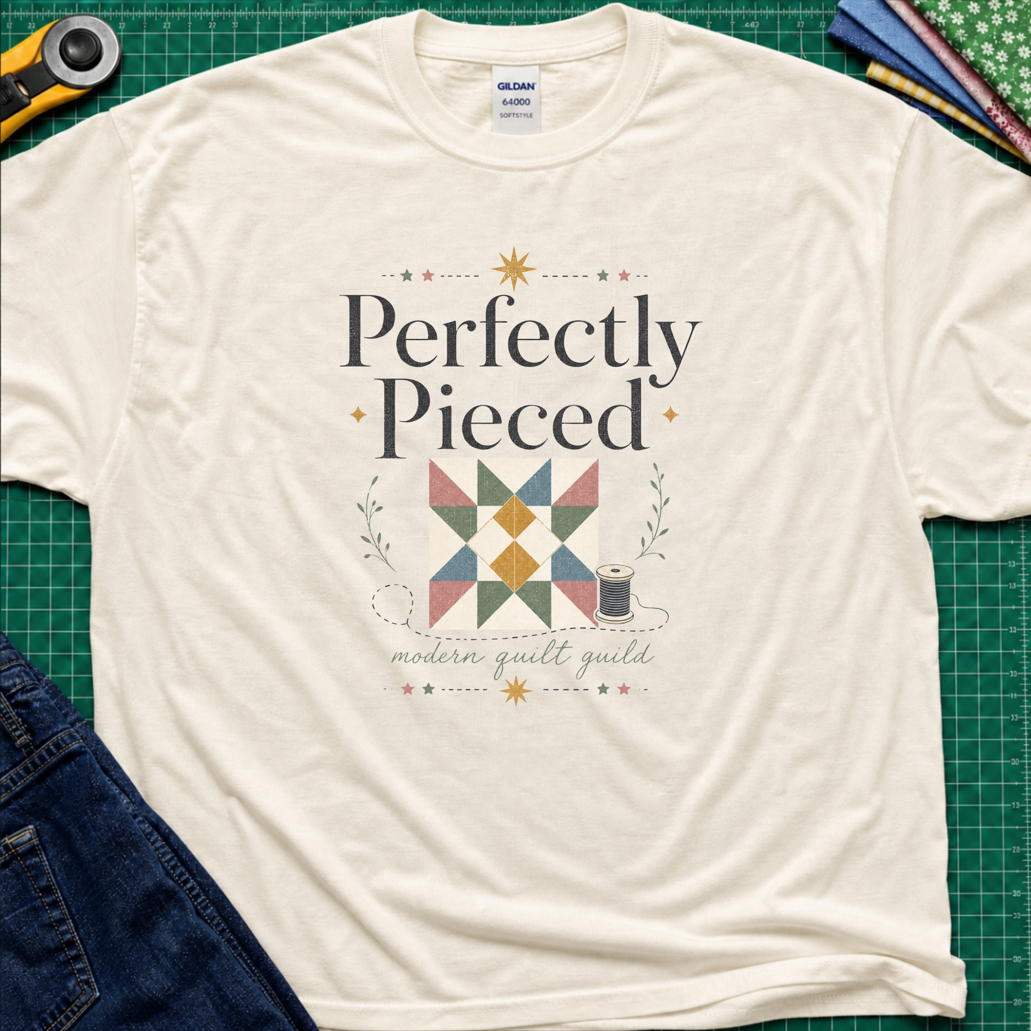 A light cream Gildan t-shirt with a quilt-themed graphic reading 'Perfectly Pieced' and 'modern quilt guild' around a patchwork block with thread spool, stars and leaves. The shirt lays flat on a green cutting mat with a rotary cutter, fabric stack and blue jeans partially visible in the corners.