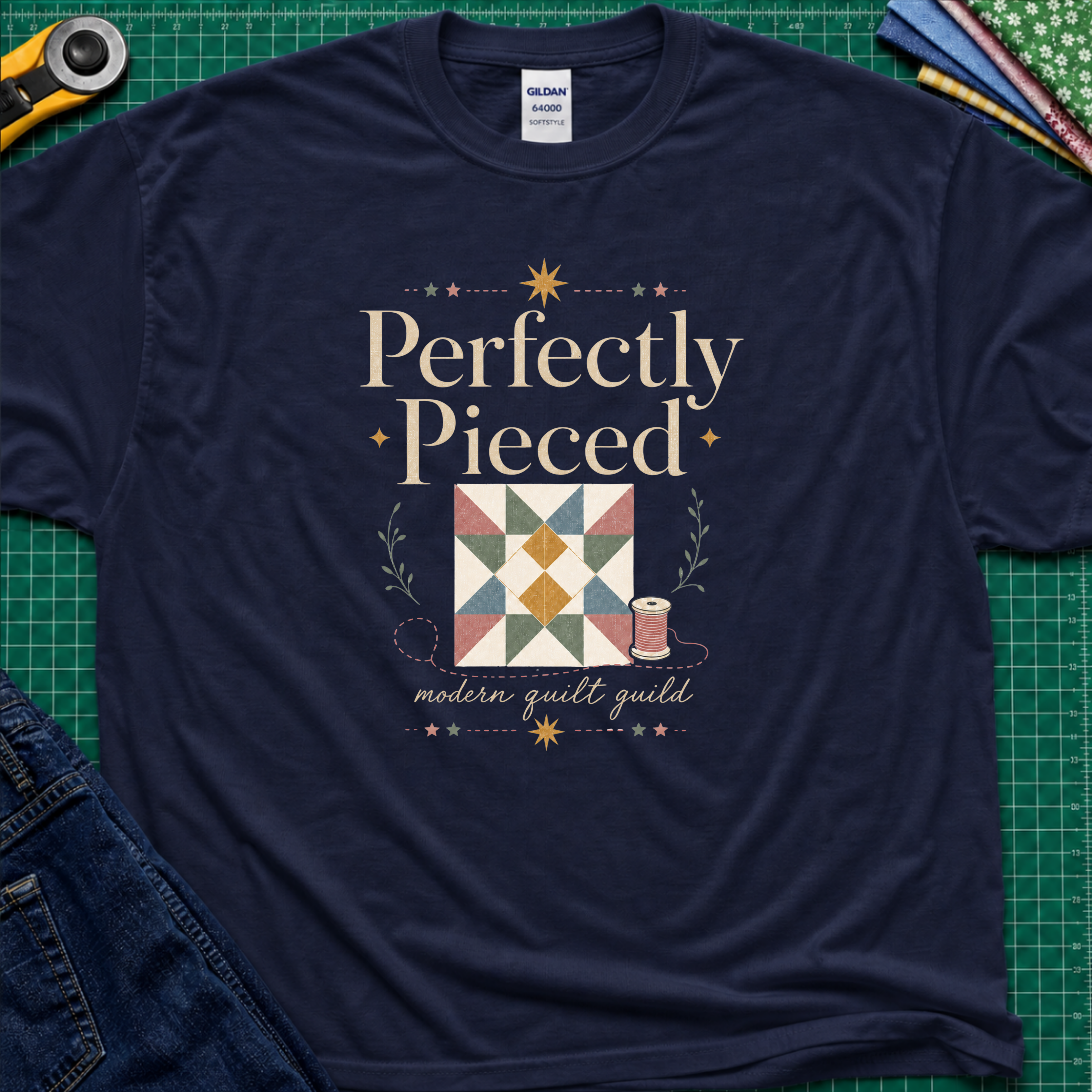 A navy blue Gildan t-shirt with a quilt-themed graphic reading 'Perfectly Pieced' and 'modern quilt guild' around a patchwork block with thread spool, stars and leaves. The shirt lays flat on a green cutting mat with a rotary cutter, fabric stack and blue jeans partially visible in the corners.