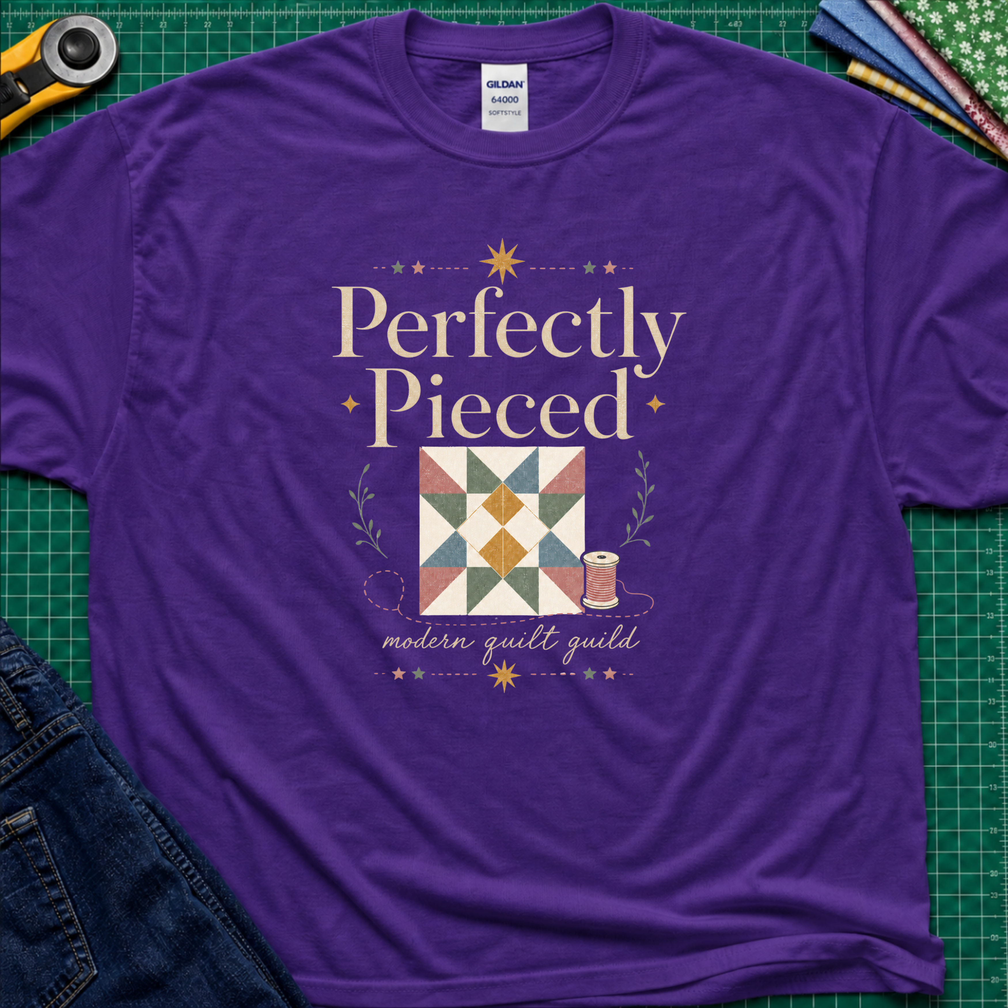 A purple Gildan t-shirt with a quilt-themed graphic reading 'Perfectly Pieced' and 'modern quilt guild' around a patchwork block with thread spool, stars and leaves. The shirt lays flat on a green cutting mat with a rotary cutter, fabric stack and blue jeans partially visible in the corners.
