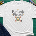 A white Gildan t-shirt with a quilt-themed graphic reading 'Perfectly Pieced' and 'modern quilt guild' around a patchwork block with thread spool, stars and leaves. The shirt lays flat on a green cutting mat with a rotary cutter, fabric stack and blue jeans partially visible in the corners.