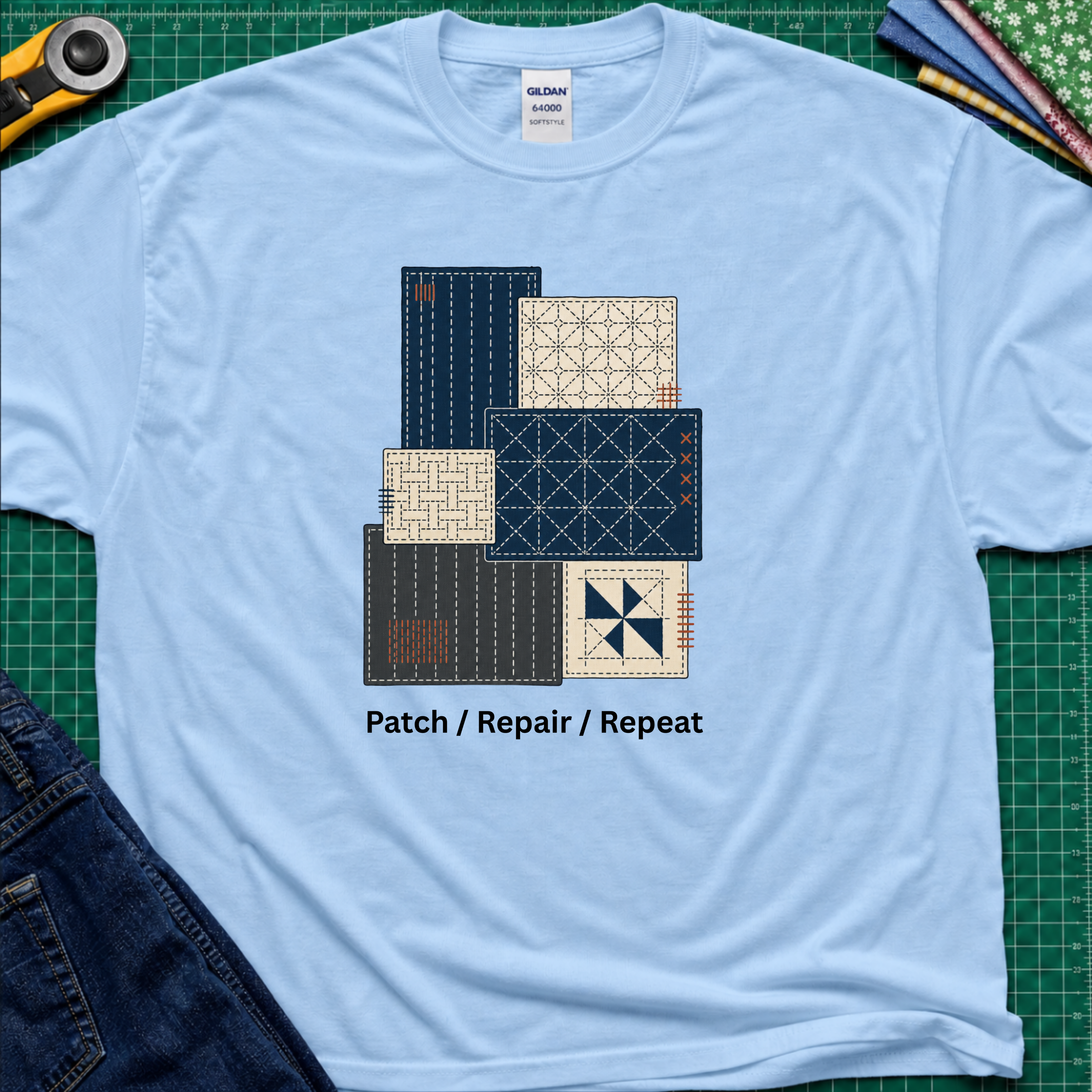 A light blue Gildan t-shirt featuring a graphic of various sashiko designs in navy, beige, and gray, with the text 'Patch / Repair / Repeat' printed below. The shirt is laid flat on a green cutting mat with a rotary cutter, fabric stack and blue jeans partially visible in the corners.
