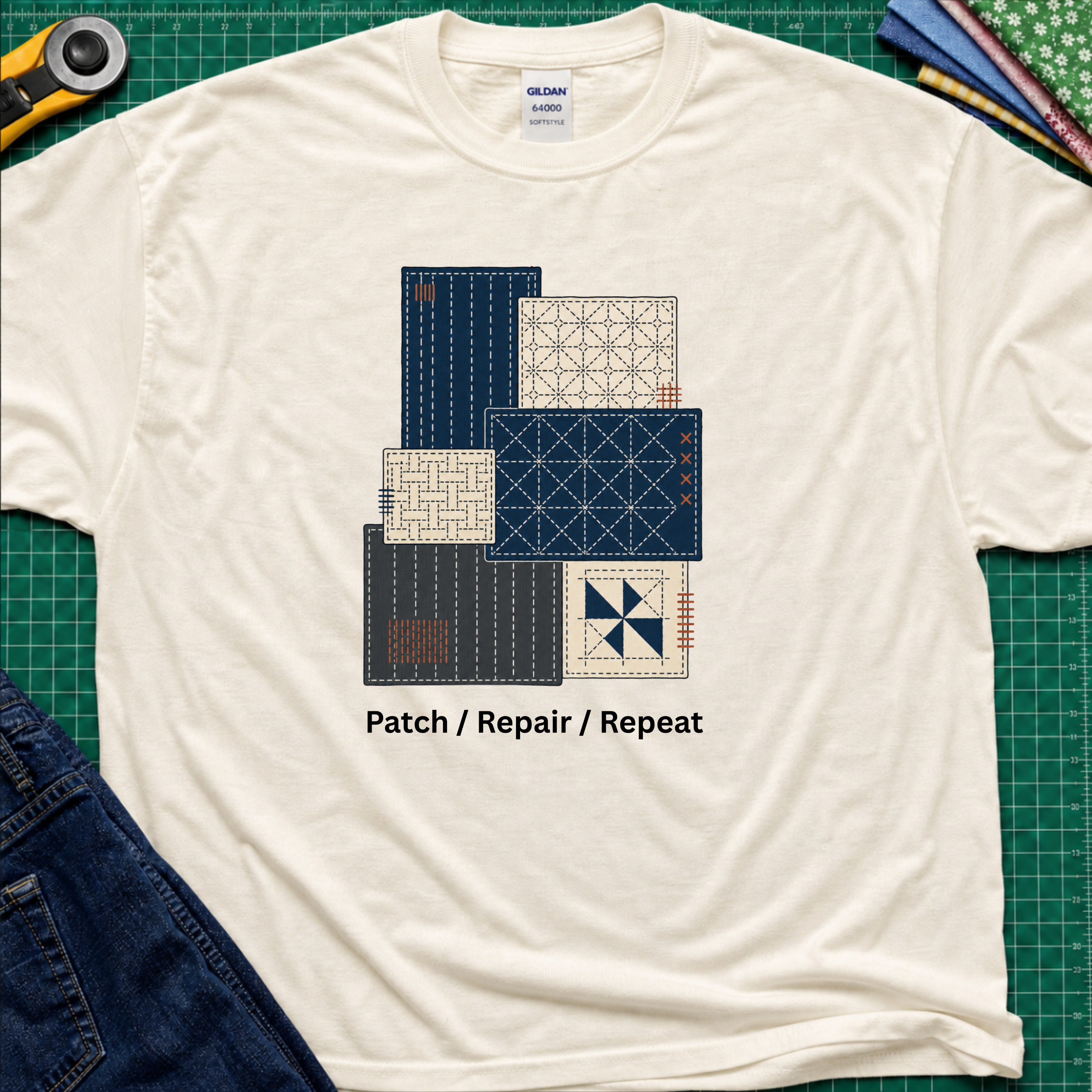 A light cream Gildan t-shirt featuring a graphic of various sashiko designs in navy, beige, and gray, with the text 'Patch / Repair / Repeat' printed below. The shirt is laid flat on a green cutting mat with a rotary cutter, fabric stack and blue jeans partially visible in the corners.
