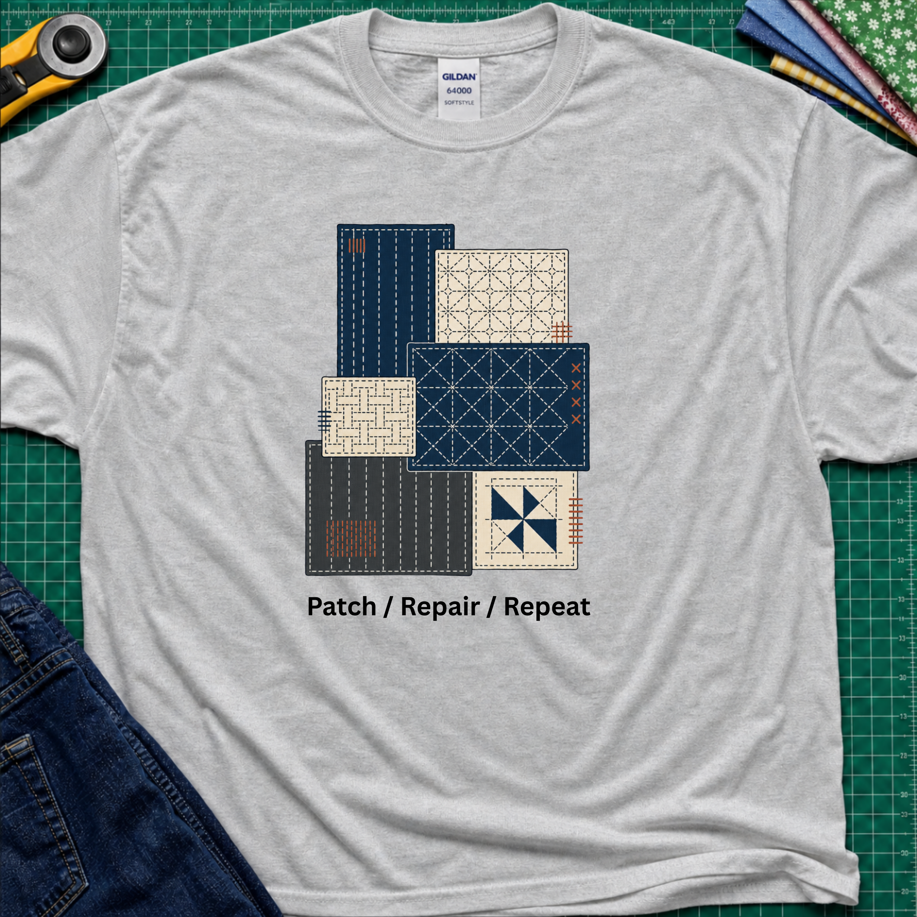 A sports grey Gildan t-shirt featuring a graphic of various sashiko designs in navy, beige, and gray, with the text 'Patch / Repair / Repeat' printed below. The shirt is laid flat on a green cutting mat with a rotary cutter, fabric stack and blue jeans partially visible in the corners.