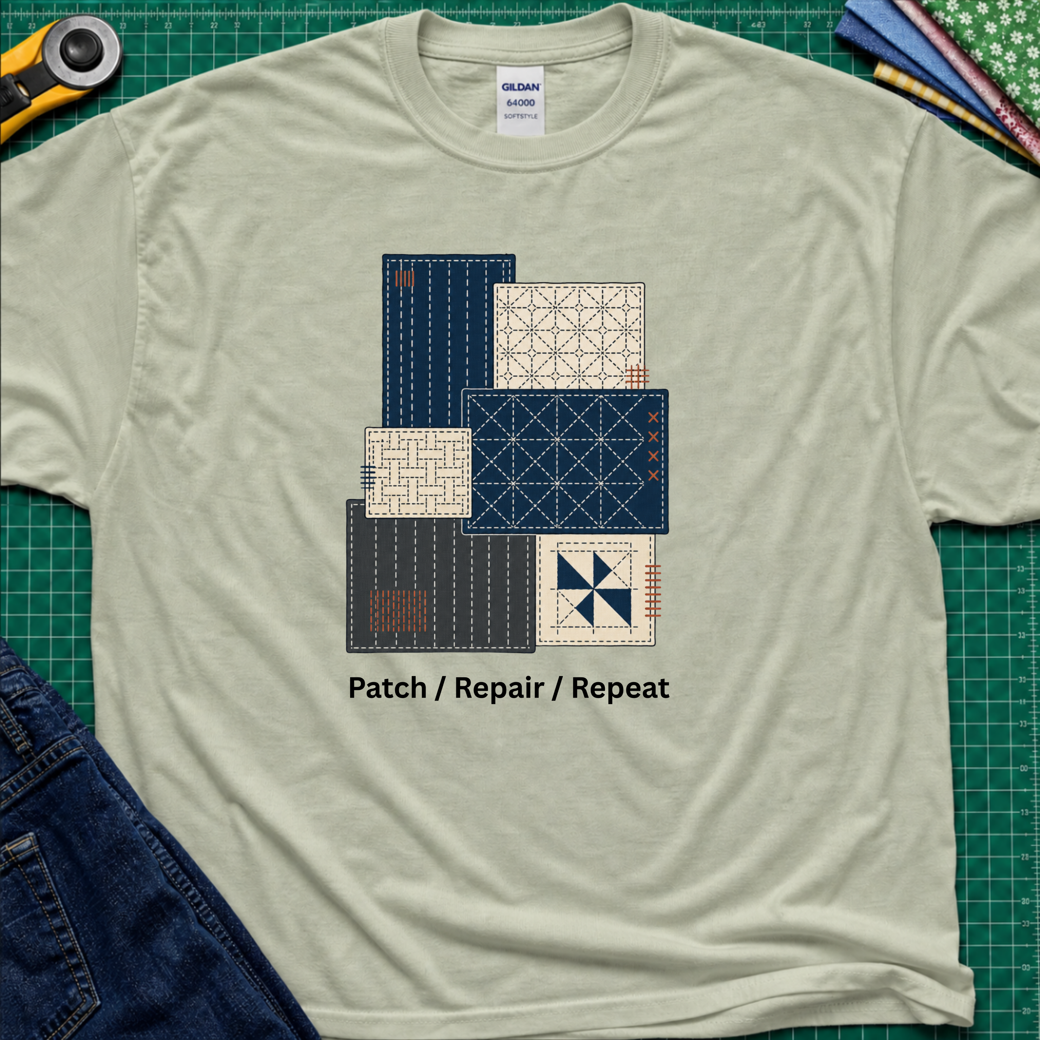 A light sage green Gildan t-shirt featuring a graphic of various sashiko designs in navy, beige, and gray, with the text 'Patch / Repair / Repeat' printed below. The shirt is laid flat on a green cutting mat with a rotary cutter, fabric stack and blue jeans partially visible in the corners.