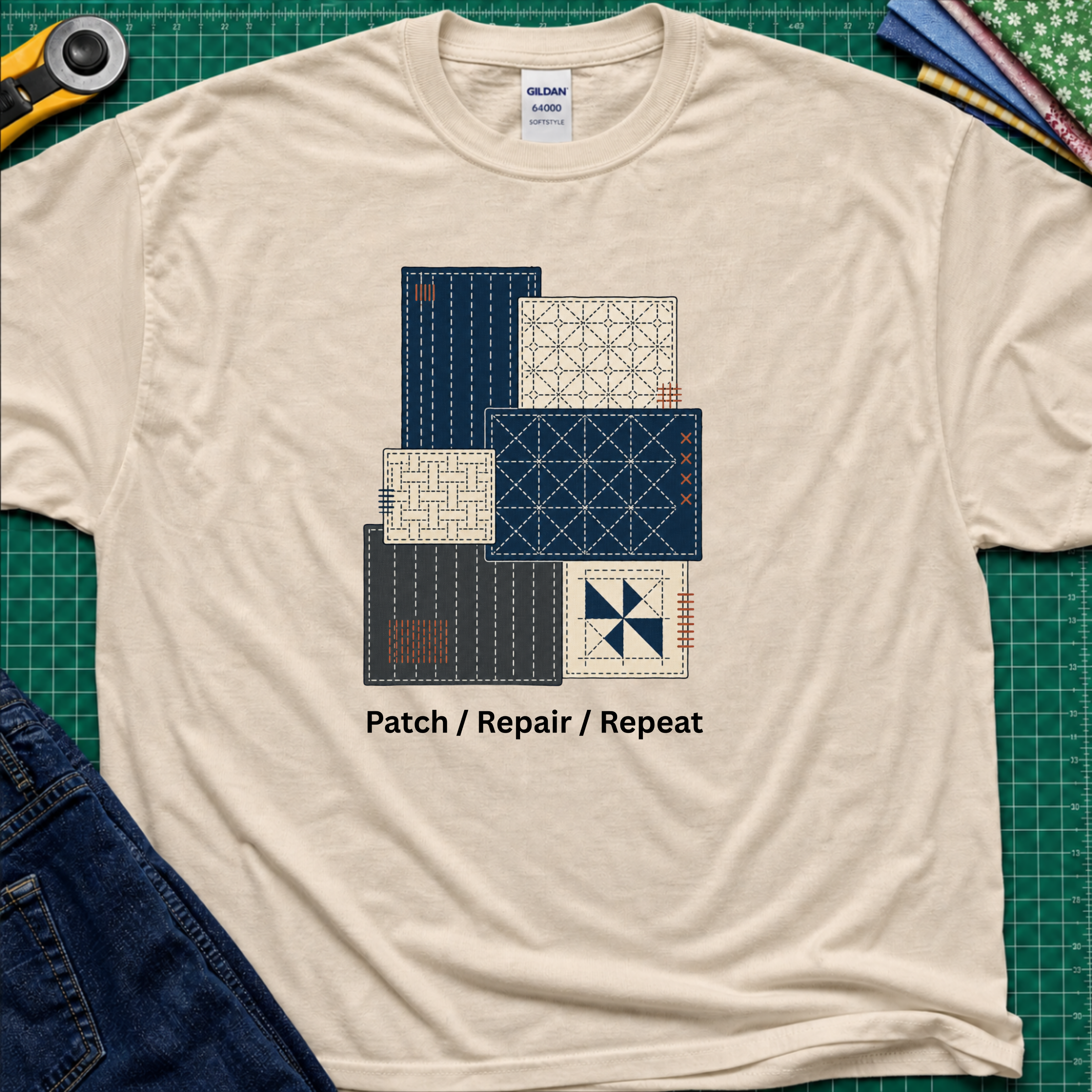 A dark cream Gildan t-shirt featuring a graphic of various sashiko designs in navy, beige, and gray, with the text 'Patch / Repair / Repeat' printed below. The shirt is laid flat on a green cutting mat with a rotary cutter, fabric stack and blue jeans partially visible in the corners.