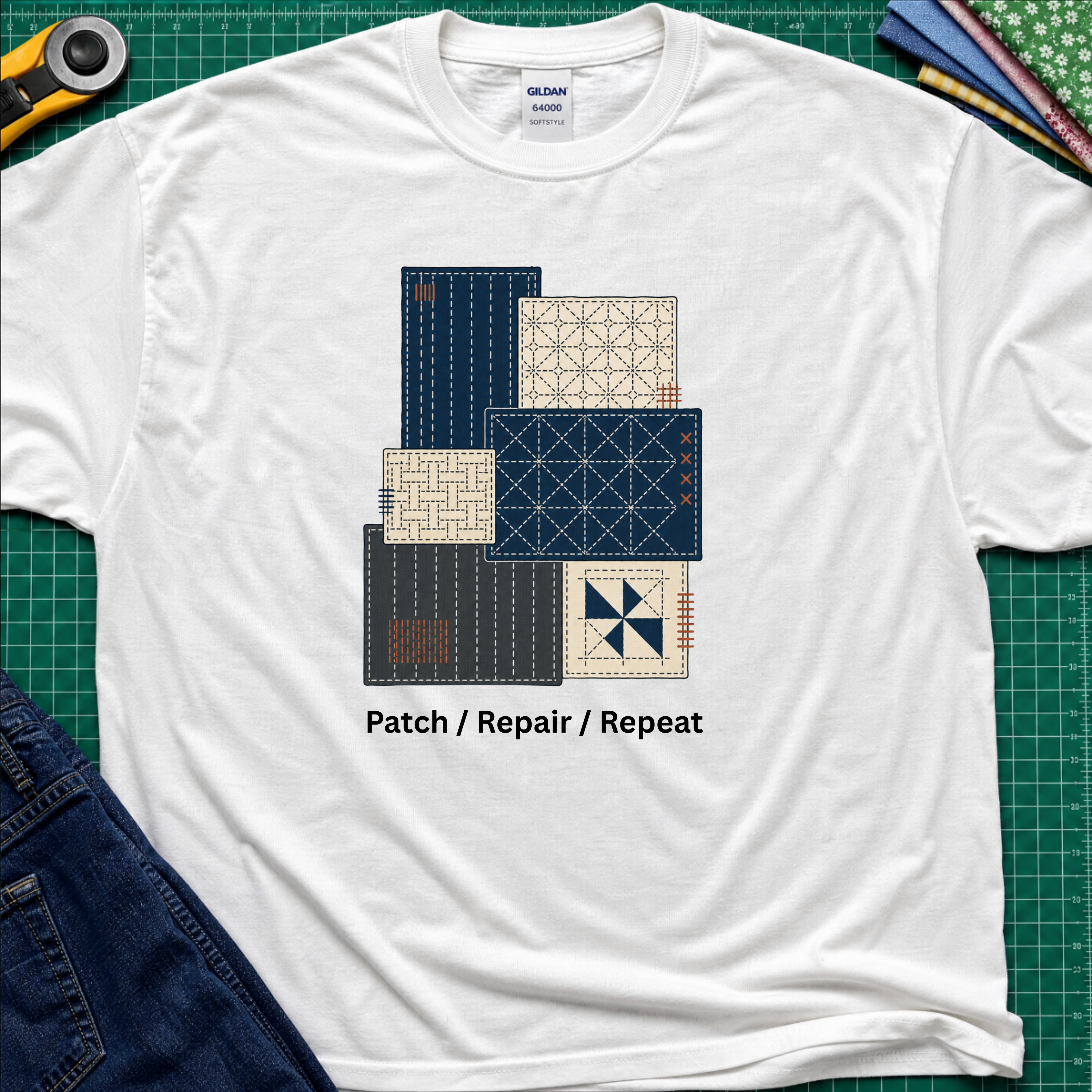 A white Gildan t-shirt featuring a graphic of various sashiko designs in navy, beige, and gray, with the text 'Patch / Repair / Repeat' printed below. The shirt is laid flat on a green cutting mat with a rotary cutter, fabric stack and blue jeans partially visible in the corners.