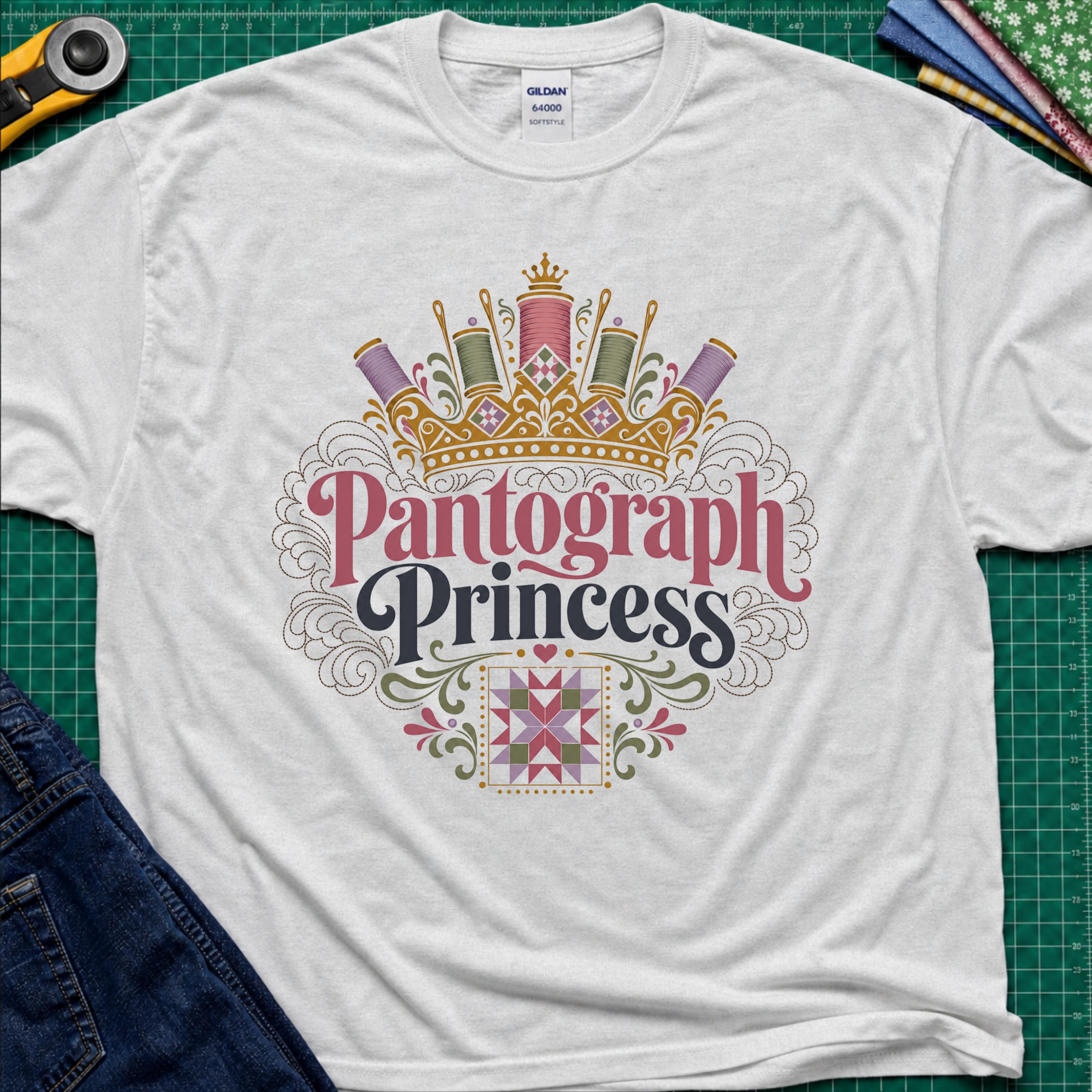 A light grey Gildan t-shirt featuring a decorative graphic that reads 'Pantograph Princess' with a crown and sewing-themed embellishments. The t-shirt lays flat on a green cutting mat with a rotary cutter, folded fabrics and blue jeans partially visible in the corners.