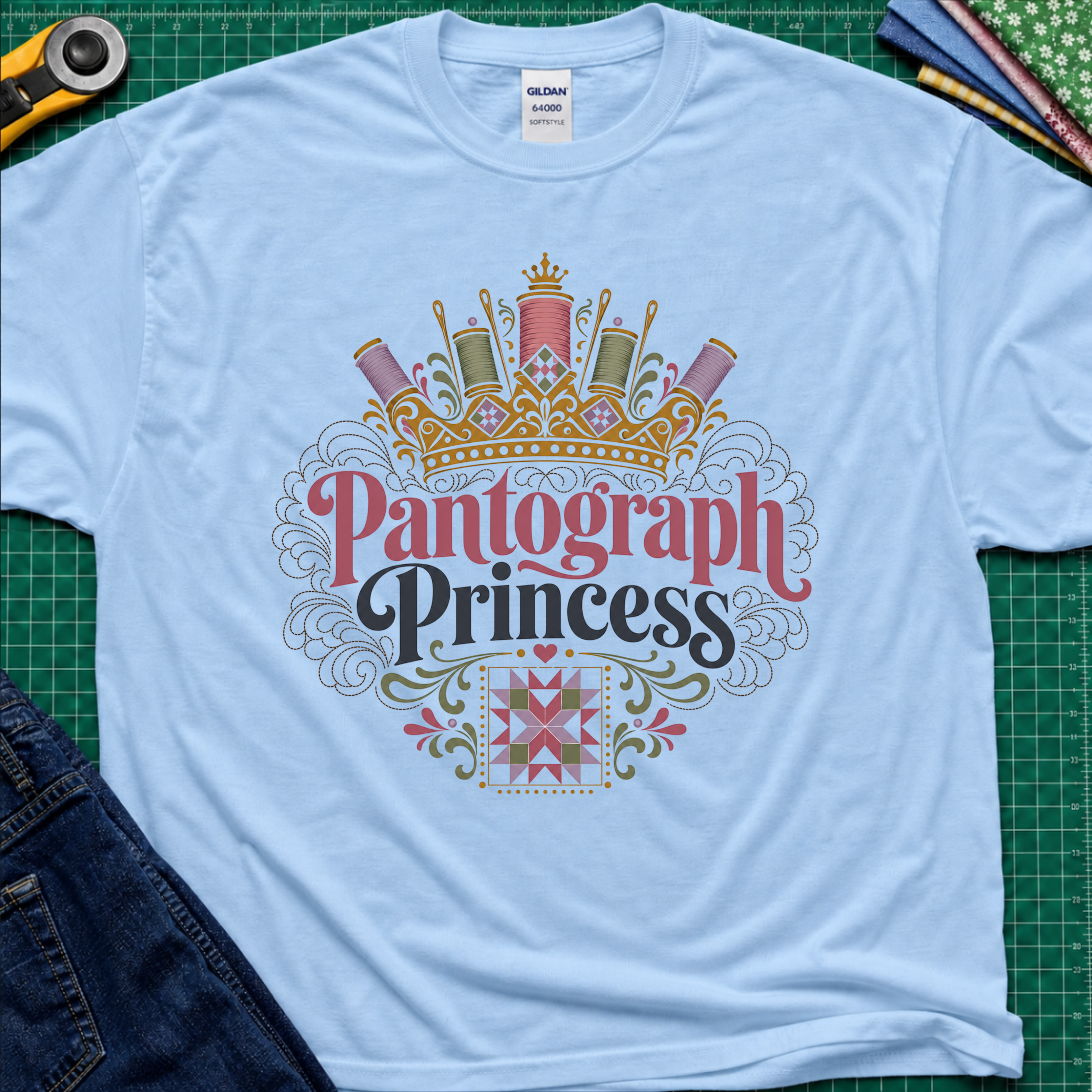 A light blue Gildan t-shirt featuring a decorative graphic that reads 'Pantograph Princess' with a crown and sewing-themed embellishments. The t-shirt lays flat on a green cutting mat with a rotary cutter, folded fabrics and blue jeans partially visible in the corners.