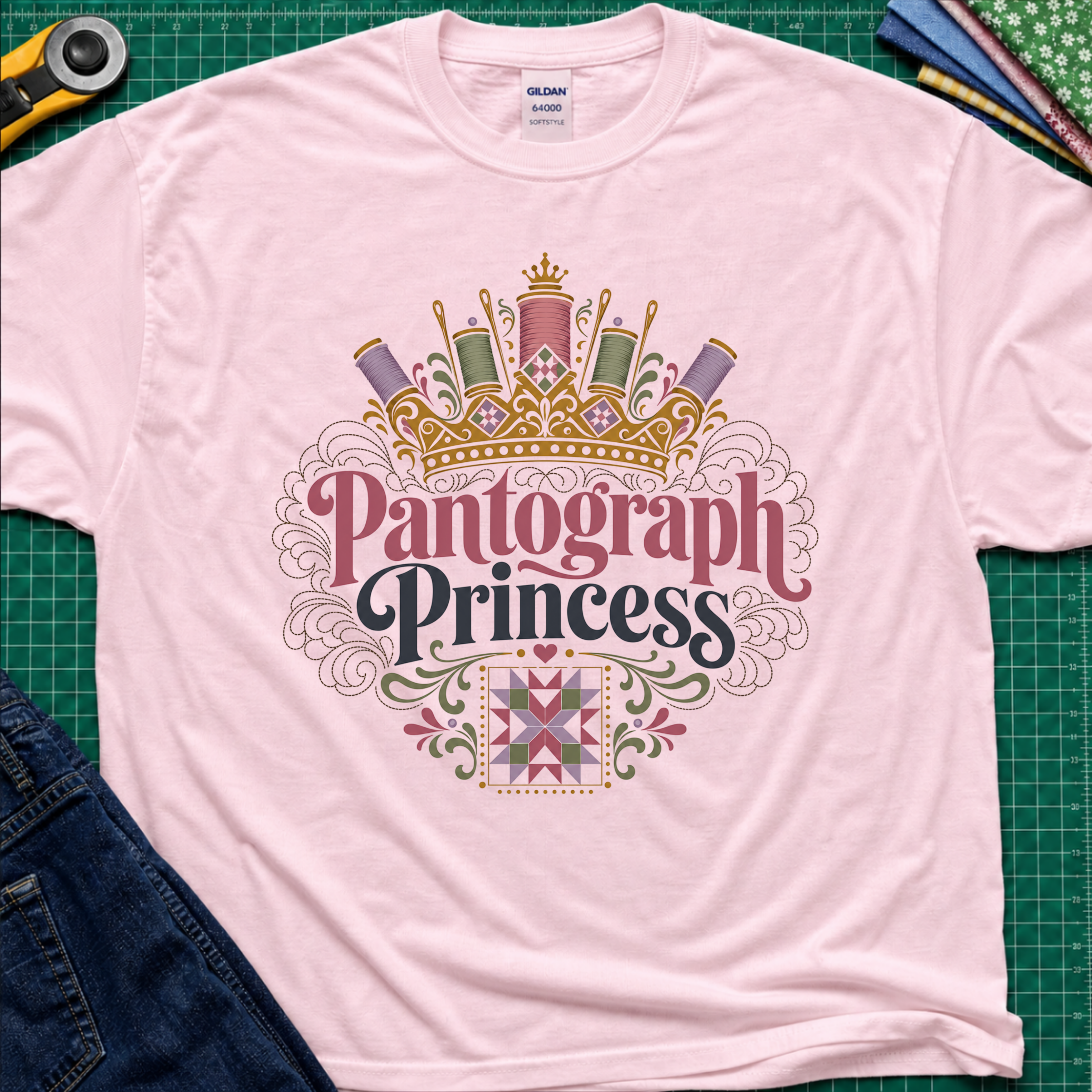 A light pink Gildan t-shirt featuring a decorative graphic that reads 'Pantograph Princess' with a crown and sewing-themed embellishments. The t-shirt lays flat on a green cutting mat with a rotary cutter, folded fabrics and blue jeans partially visible in the corners.