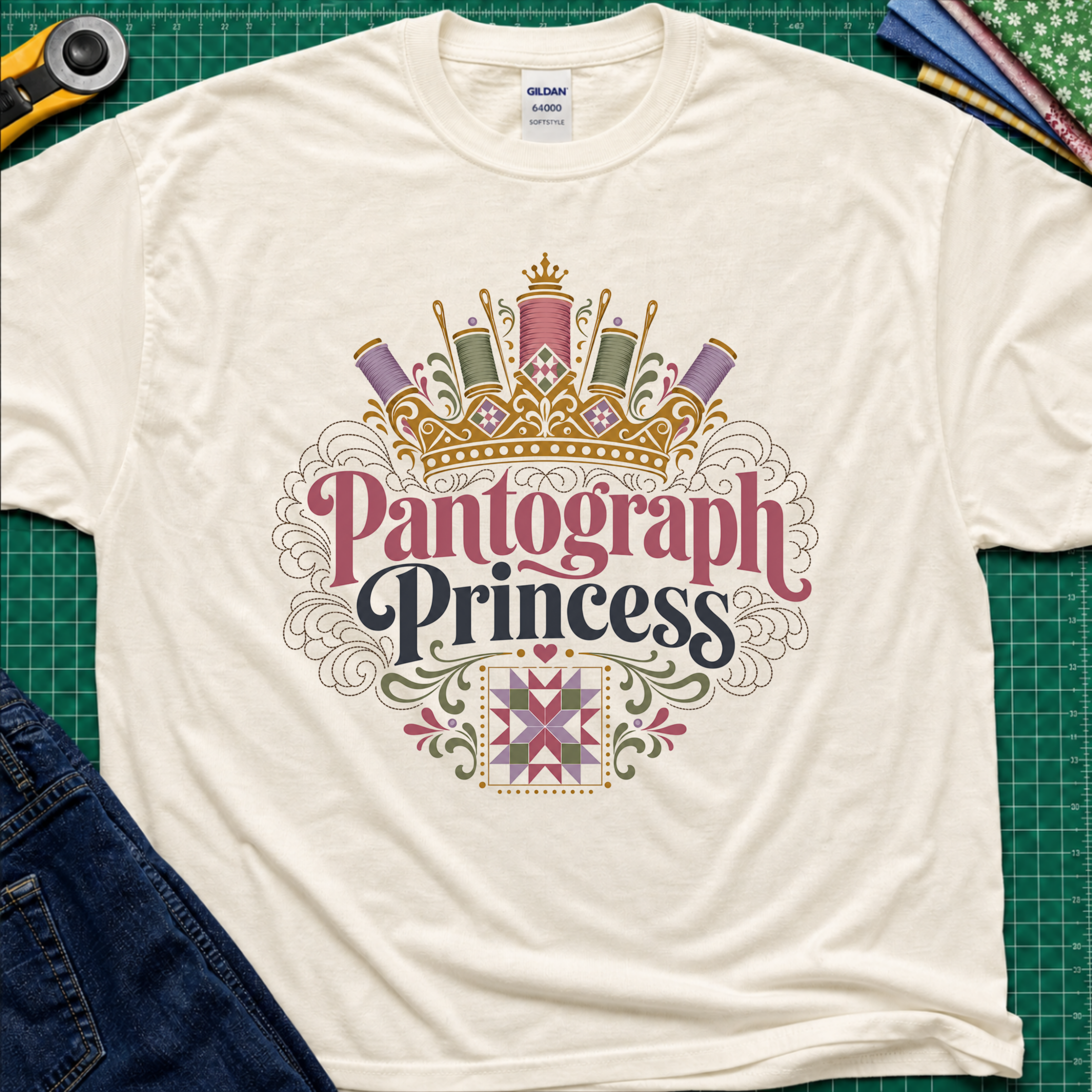 A light cream Gildan t-shirt featuring a decorative graphic that reads 'Pantograph Princess' with a crown and sewing-themed embellishments. The t-shirt lays flat on a green cutting mat with a rotary cutter, folded fabrics and blue jeans partially visible in the corners.