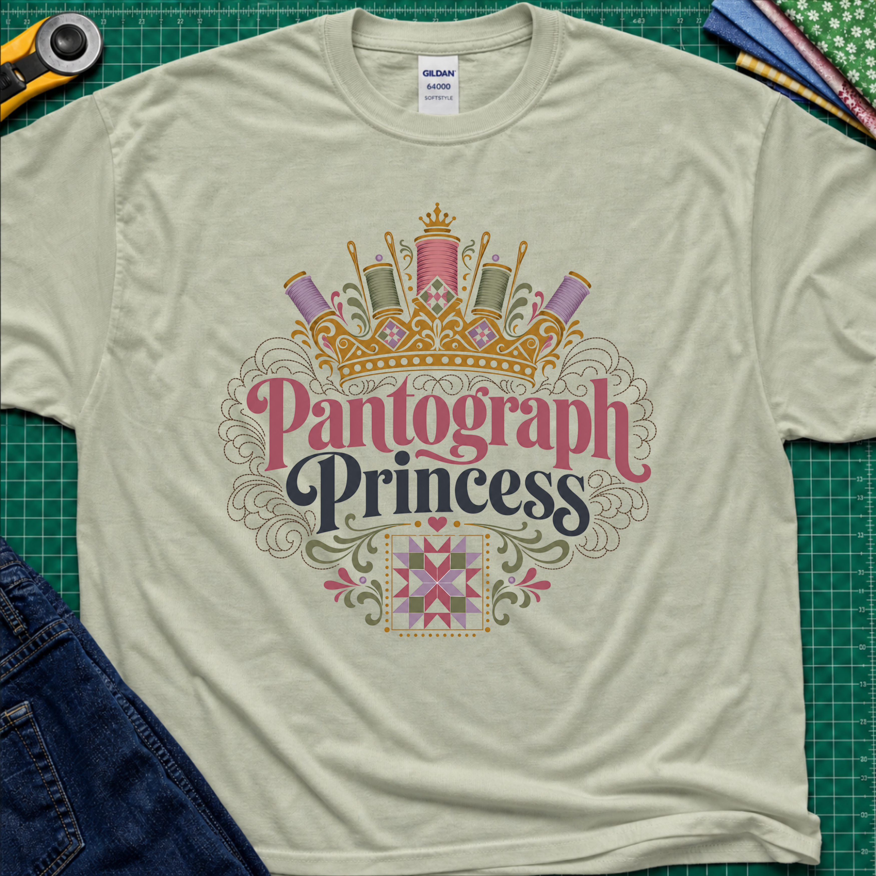 A light sage green Gildan t-shirt featuring a decorative graphic that reads 'Pantograph Princess' with a crown and sewing-themed embellishments. The t-shirt lays flat on a green cutting mat with a rotary cutter, folded fabrics and blue jeans partially visible in the corners.