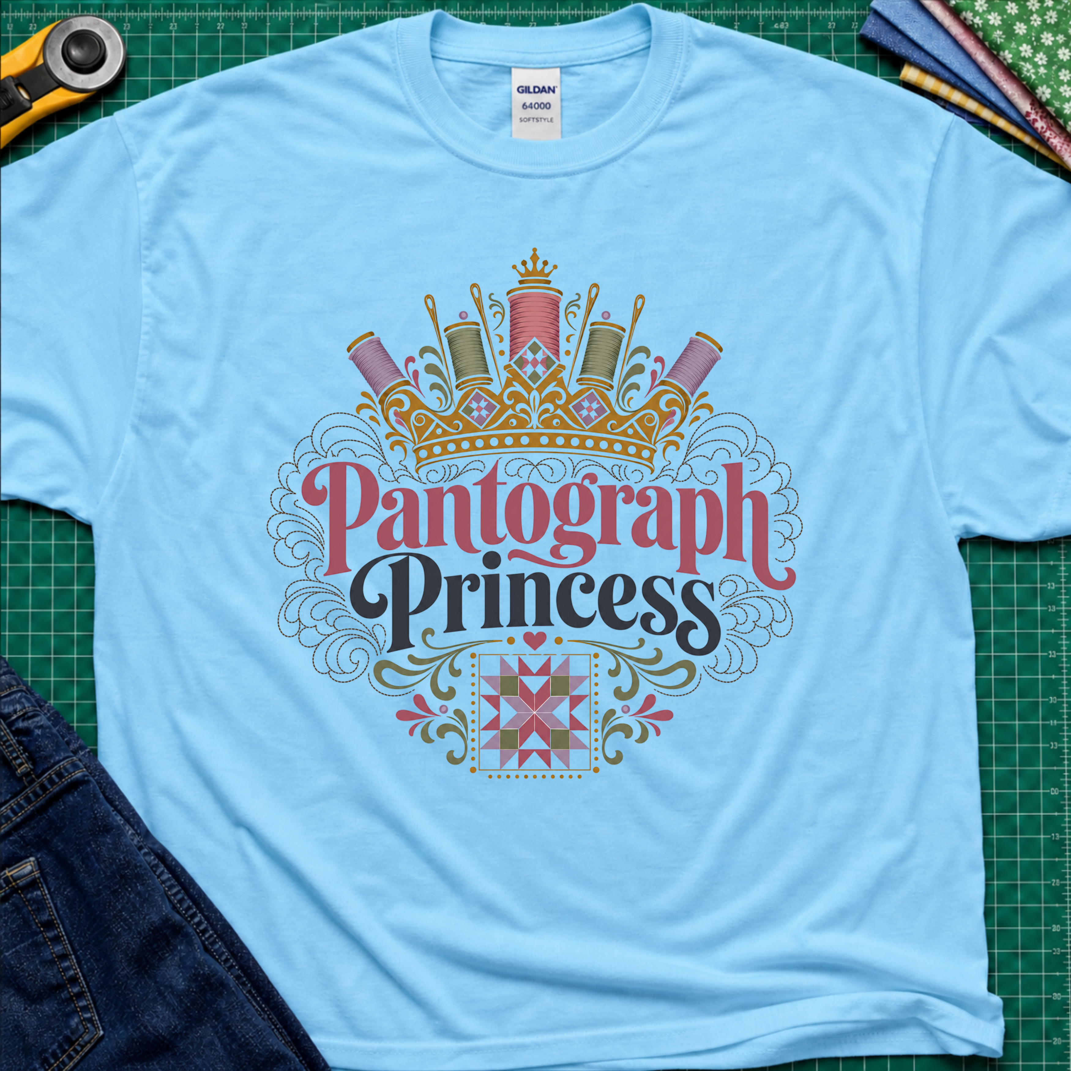 A sky blue Gildan t-shirt featuring a decorative graphic that reads 'Pantograph Princess' with a crown and sewing-themed embellishments. The t-shirt lays flat on a green cutting mat with a rotary cutter, folded fabrics and blue jeans partially visible in the corners.