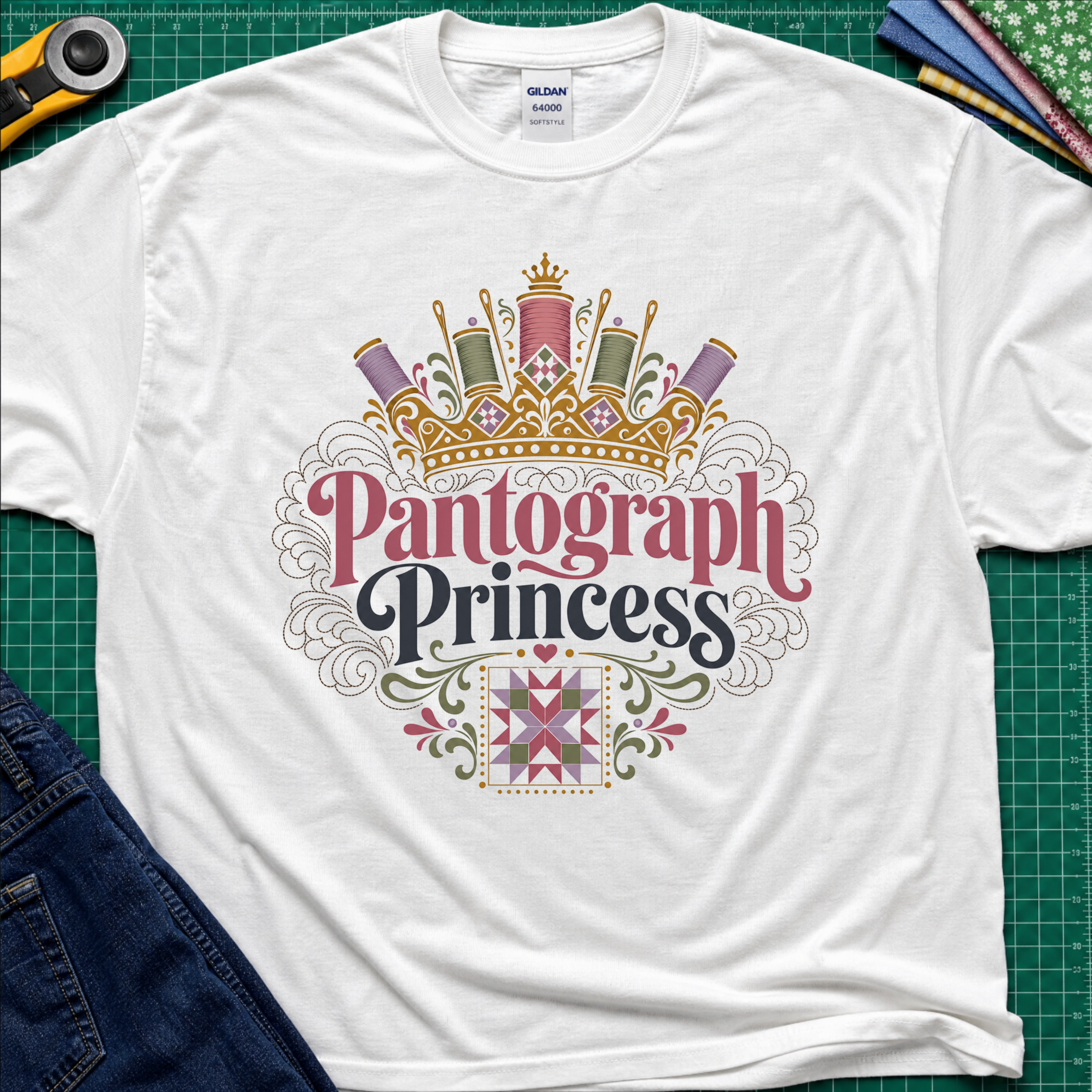 A white Gildan t-shirt featuring a decorative graphic that reads 'Pantograph Princess' with a crown and sewing-themed embellishments. The t-shirt lays flat on a green cutting mat with a rotary cutter, folded fabrics and blue jeans partially visible in the corners.