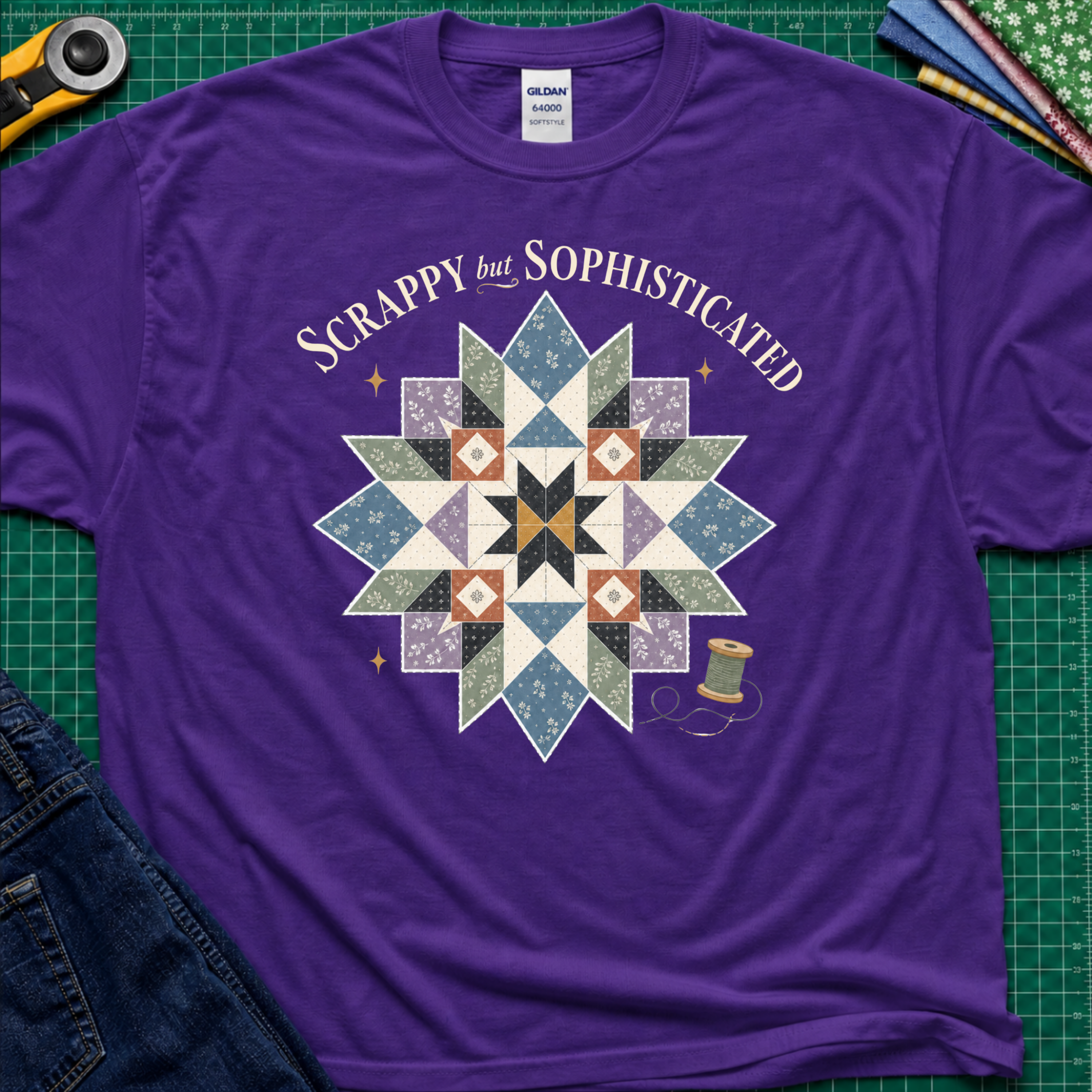 A purple Gildan t-shirt with a colorful quilted starburst graphic and the text 'Scrappy but Sophisticated' printed on the front, styled on a green cutting mat with sewing tools and denim.