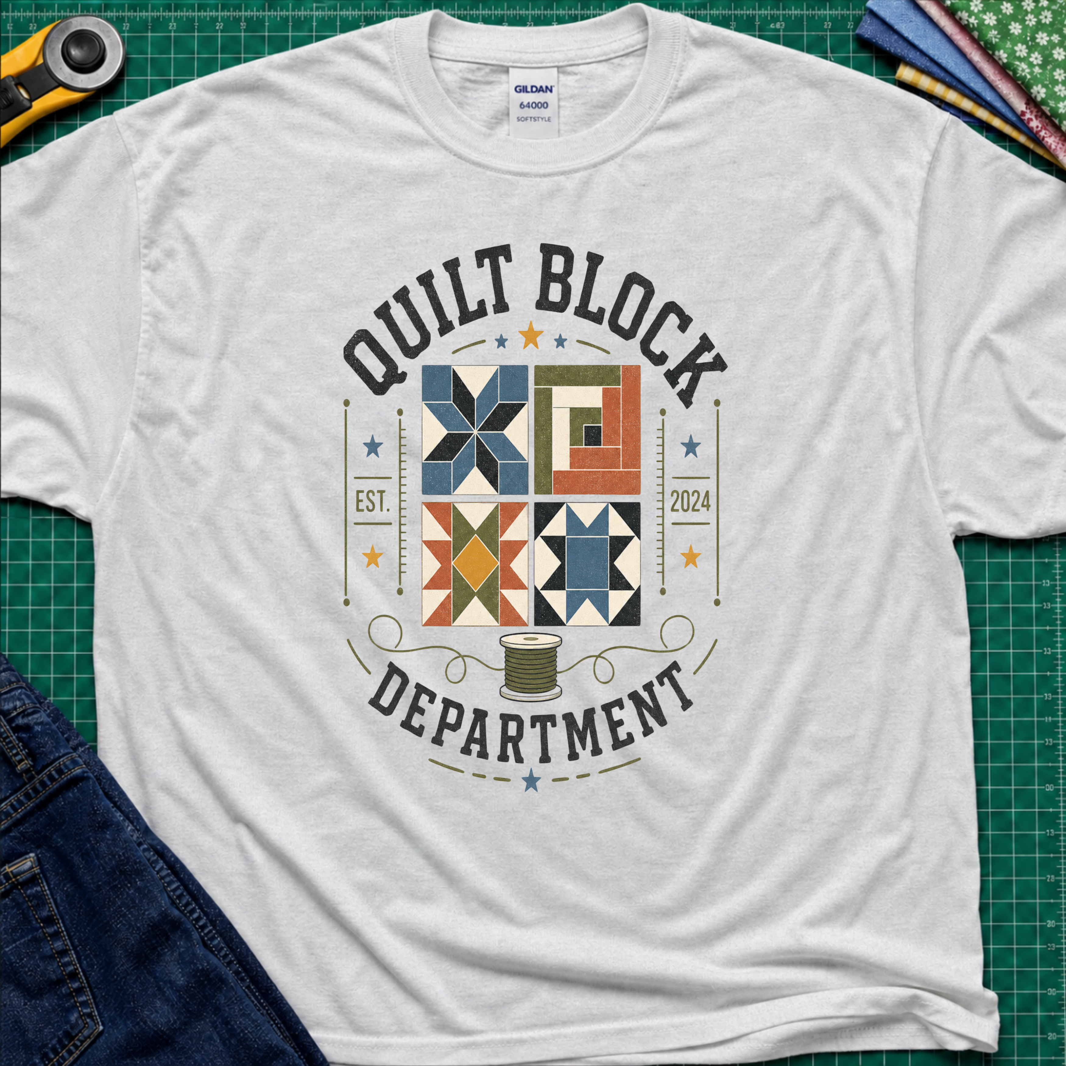 A light grey Gildan t-shirt with a colorful quilt-themed graphic reading 'QUILT BLOCK DEPARTMENT EST. 2024', surrounding a set of four blocks and a spool of thread. The shirt lays flat on a green cutting mat with a rotary cutter, fabric stack and jeans partially visible around it.