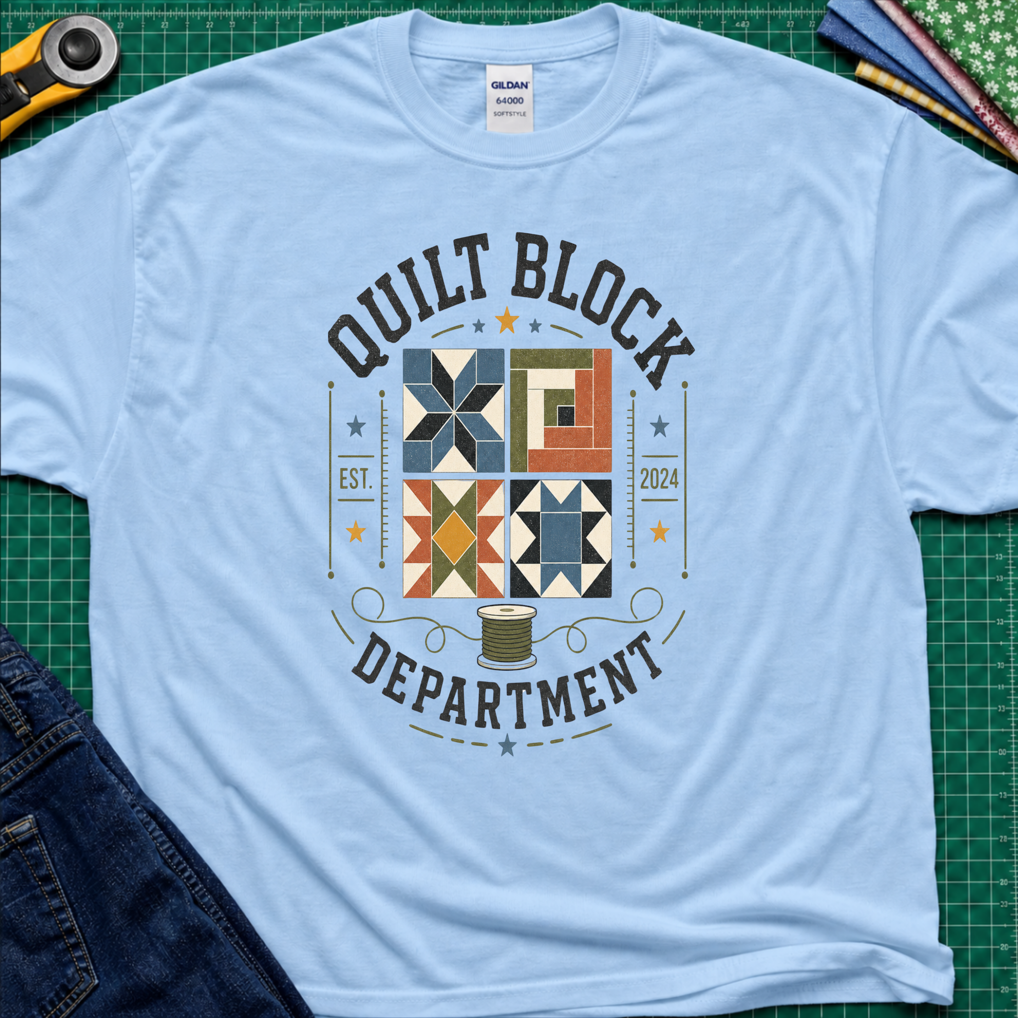 A light blue Gildan t-shirt with a colorful quilt-themed graphic reading 'QUILT BLOCK DEPARTMENT EST. 2024', surrounding a set of four blocks and a spool of thread. The shirt lays flat on a green cutting mat with a rotary cutter, fabric stack and jeans partially visible around it.