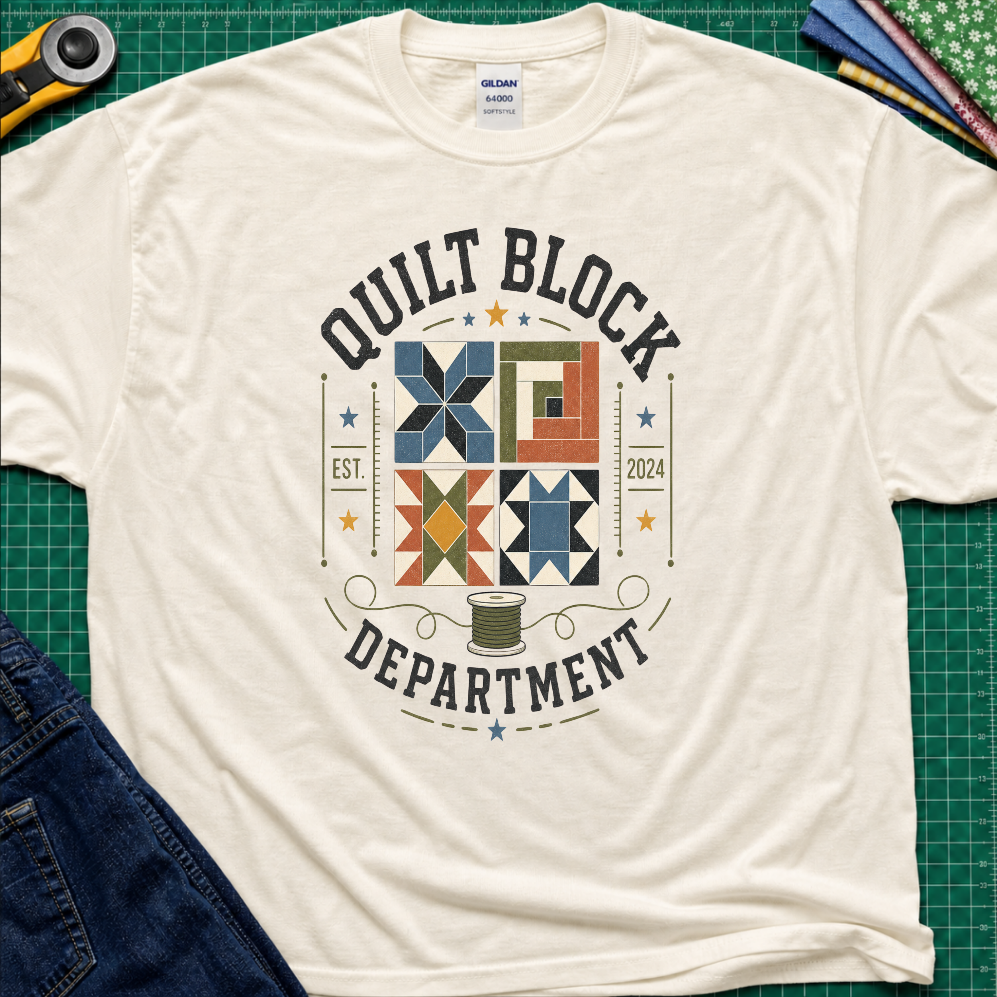 A light cream Gildan t-shirt with a colorful quilt-themed graphic reading 'QUILT BLOCK DEPARTMENT EST. 2024', surrounding a set of four blocks and a spool of thread. The shirt lays flat on a green cutting mat with a rotary cutter, fabric stack and jeans partially visible around it.