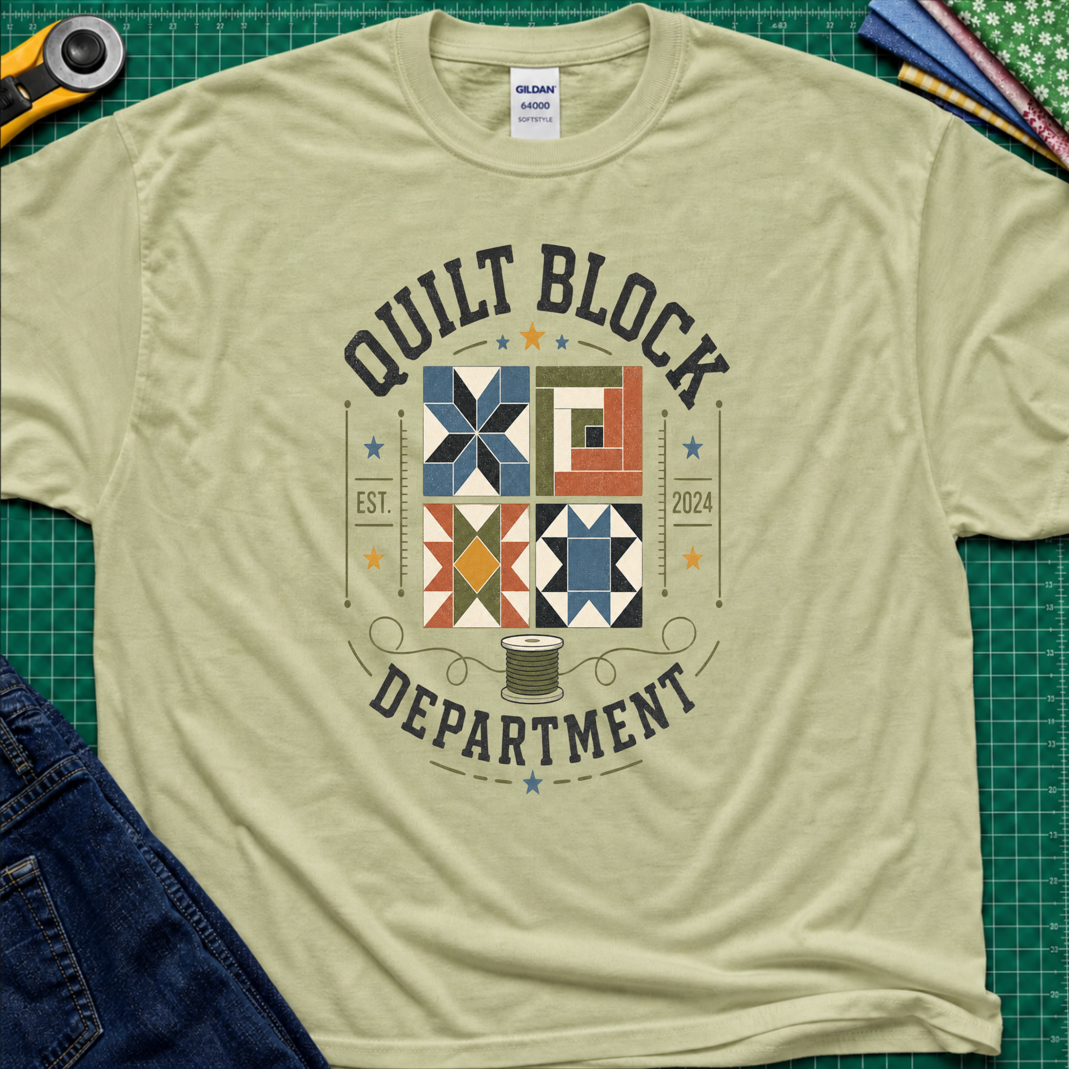 A pistachio green Gildan t-shirt with a colorful quilt-themed graphic reading 'QUILT BLOCK DEPARTMENT EST. 2024', surrounding a set of four blocks and a spool of thread. The shirt lays flat on a green cutting mat with a rotary cutter, fabric stack and jeans partially visible around it.