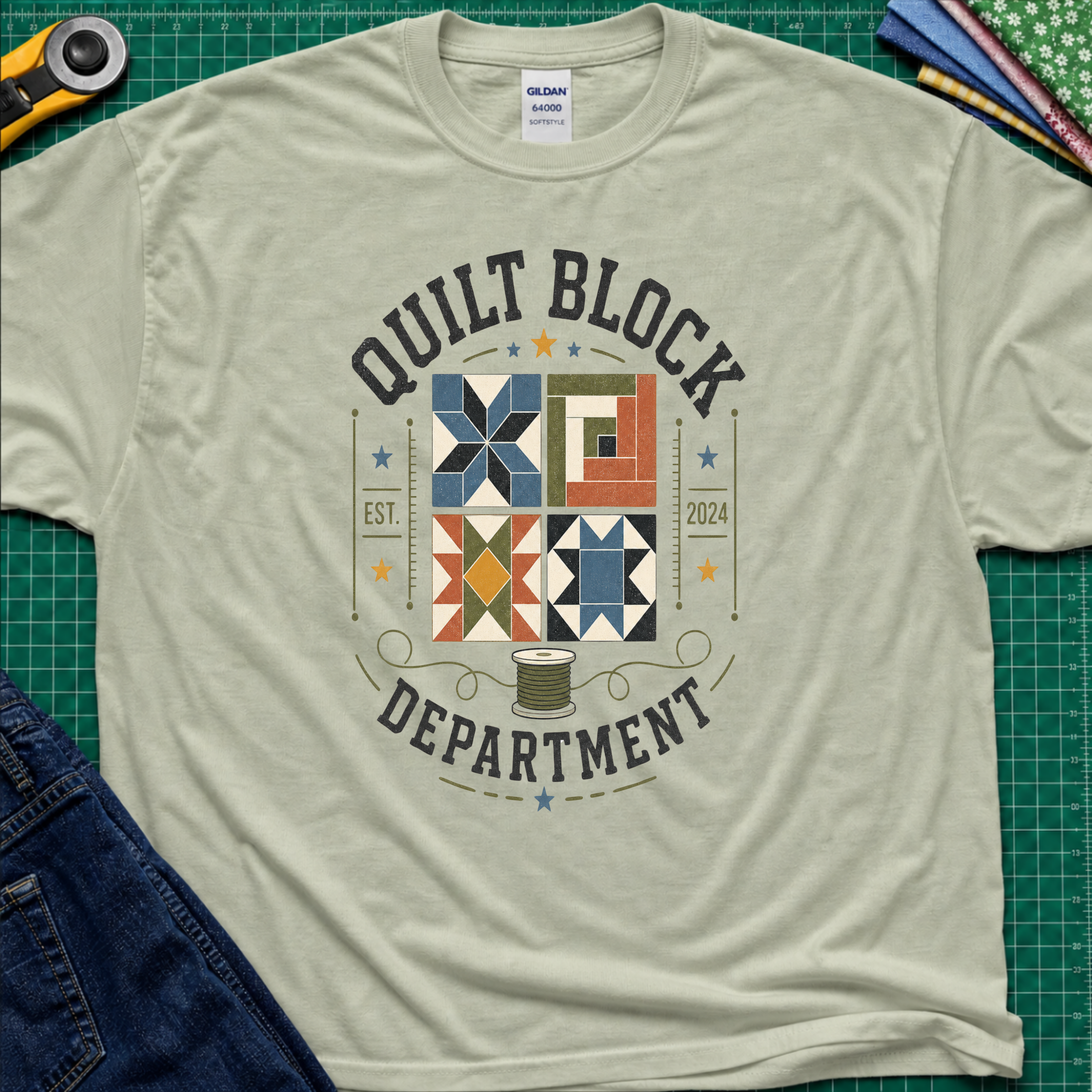 A light sage green Gildan t-shirt with a colorful quilt-themed graphic reading 'QUILT BLOCK DEPARTMENT EST. 2024', surrounding a set of four blocks and a spool of thread. The shirt lays flat on a green cutting mat with a rotary cutter, fabric stack and jeans partially visible around it.