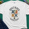 A white Gildan t-shirt with a colorful quilt-themed graphic reading 'QUILT BLOCK DEPARTMENT EST. 2024', surrounding a set of four blocks and a spool of thread. The shirt lays flat on a green cutting mat with a rotary cutter, fabric stack and jeans partially visible around it.
